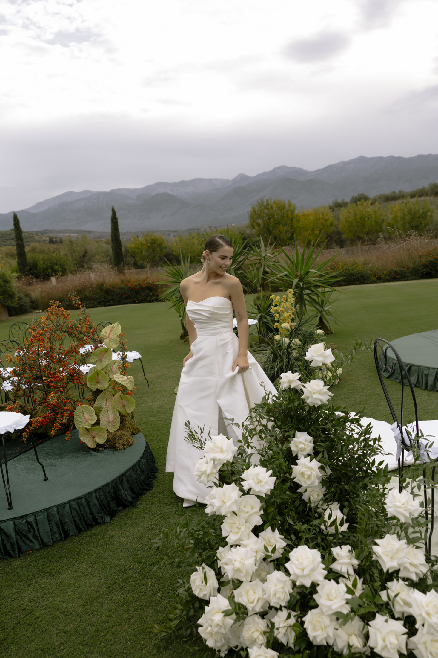 A bride in a strapless white satin gown with a softly draped bodice walks across a lawn at Roxani Estate in Greece, where a long, trailing line of white garden roses and green foliage spills along the grass beside her. Behind her, cream and green orchids rise among palms, while on the left a round green-draped platform holds a cluster of orange berried shrubs and pale green anthurium. Soft, even daylight gives the scene a calm, airy, elegant mood, with the wedding chairs dressed in white ribbons.
