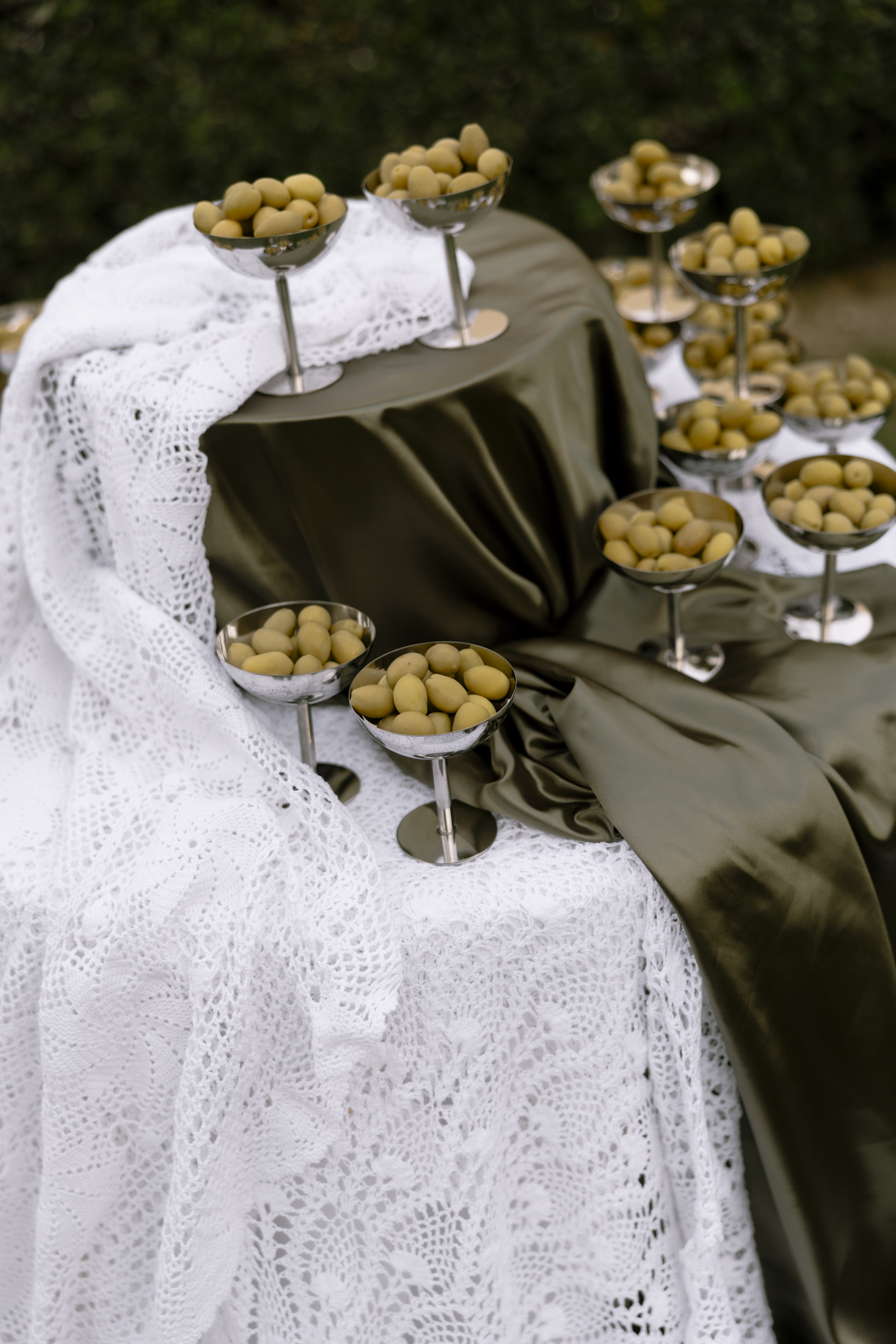 Silver footed coupe glasses, each heaped with pale green olives, stand on a table draped in deep olive-green satin and a white crocheted lace cloth, set outdoors at Roxani Estate in Greece. Olives on pedestal dishes are a striking, natural touch for a wedding table display, and the soft garden greenery behind them echoes the palette. The light is soft and even, giving the scene a calm, earthy mood.