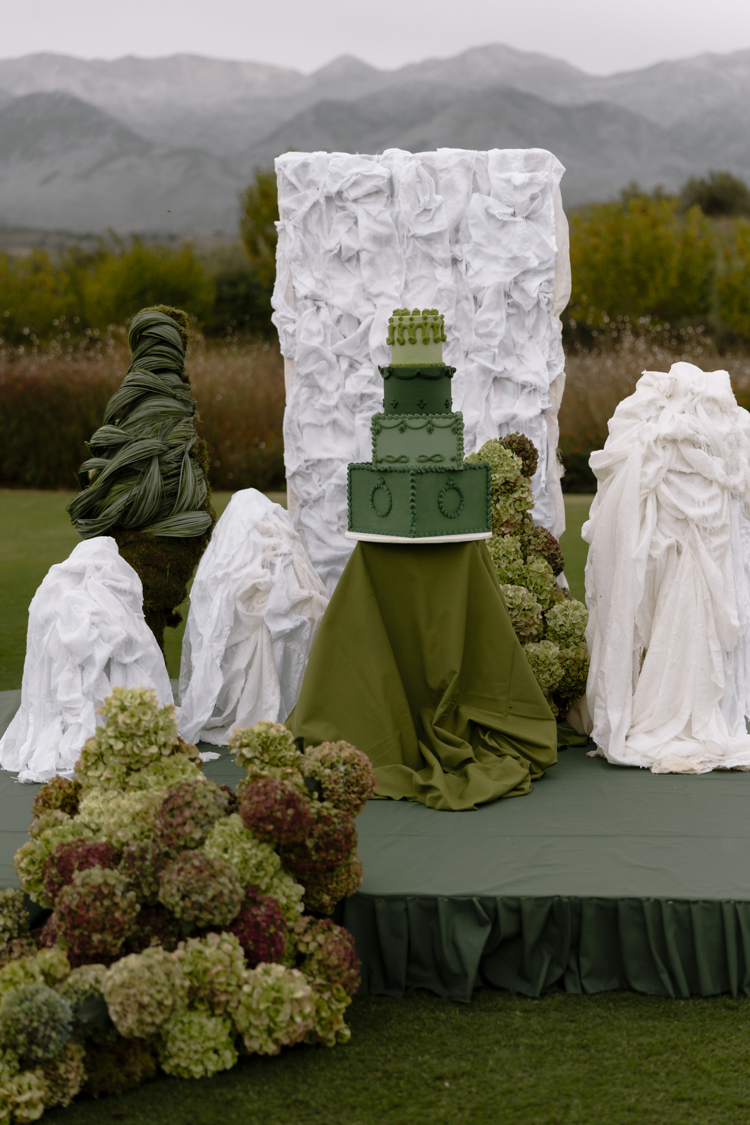 A four-tier wedding cake in shades of green, from deep forest to pale sage, stands on an olive-green draped pedestal at Roxani Estate in Greece. Around it, green and cream hydrangeas, a twisted tower of glossy leaves, and sculptural white fabric forms rise from the lawn, with a tall white draped column behind. The soft outdoor light and muted greens give the scene a calm, romantic feel.