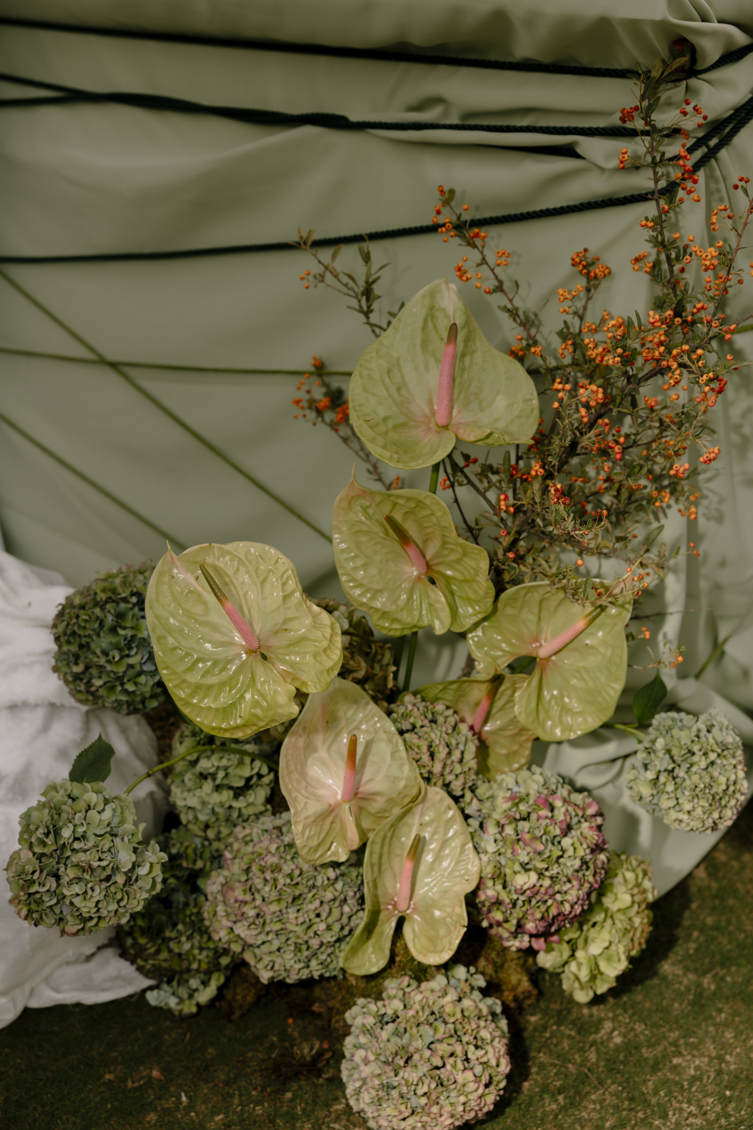Pale green anthurium blooms, their glossy heart-shaped spathes curving around pink spadices, rise through a cluster of softly faded green hydrangea heads. Sprigs of tiny orange pyracantha berries add a warm accent against a sage-green draped backdrop. These are wedding flowers from The Green Edit story at Roxani Estate in Greece, arranged in a tonal, nature-inspired display.