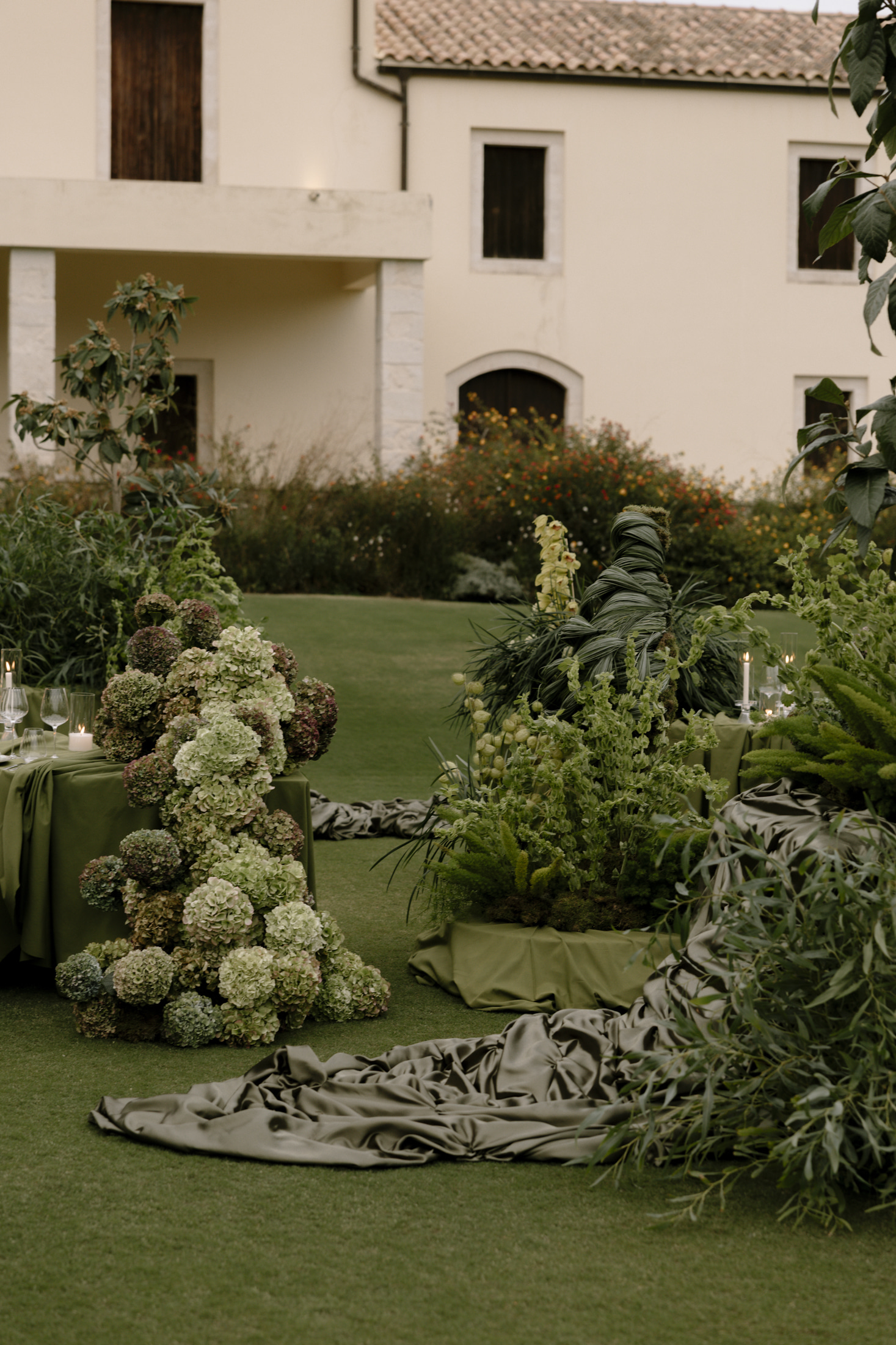 Wedding table flowers set on a lawn in front of a cream stone-trimmed villa at Roxani Estate in Greece: a low cluster of pale green and burgundy hydrangea heads sits on a moss-green tablecloth beside a candle and wine glasses. Behind it, tall spikes of pale green orchids, arching grasses and a twisted dark foliage sculpture rise among trailing green stems, with a bed of orange and rust blooms in the background.