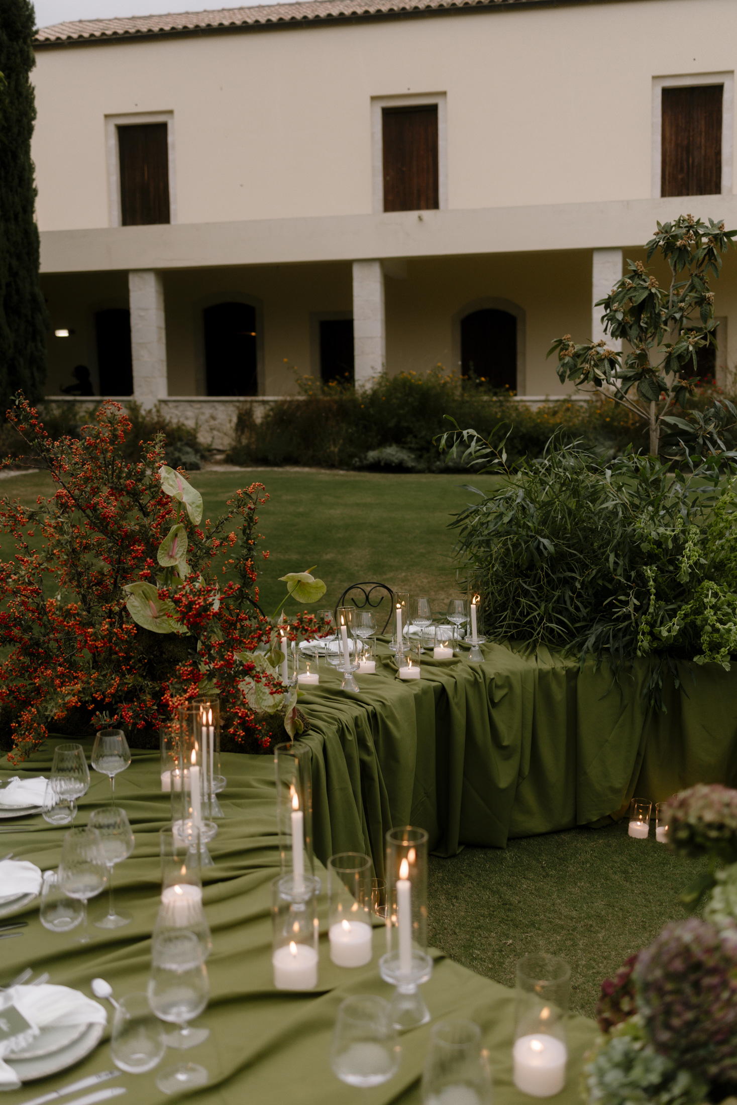 A long wedding table draped in olive-green cloth sits on a lawn at Roxani Estate in Greece, lit by tall taper and tealight candles among clear glassware. On the left, a bush of red-orange berries is paired with pale green anthurium blooms, while willowy green foliage fills the right side. Behind the table, a cream two-storey building with arched doorways and a lawn in soft evening light creates a calm, understated mood.