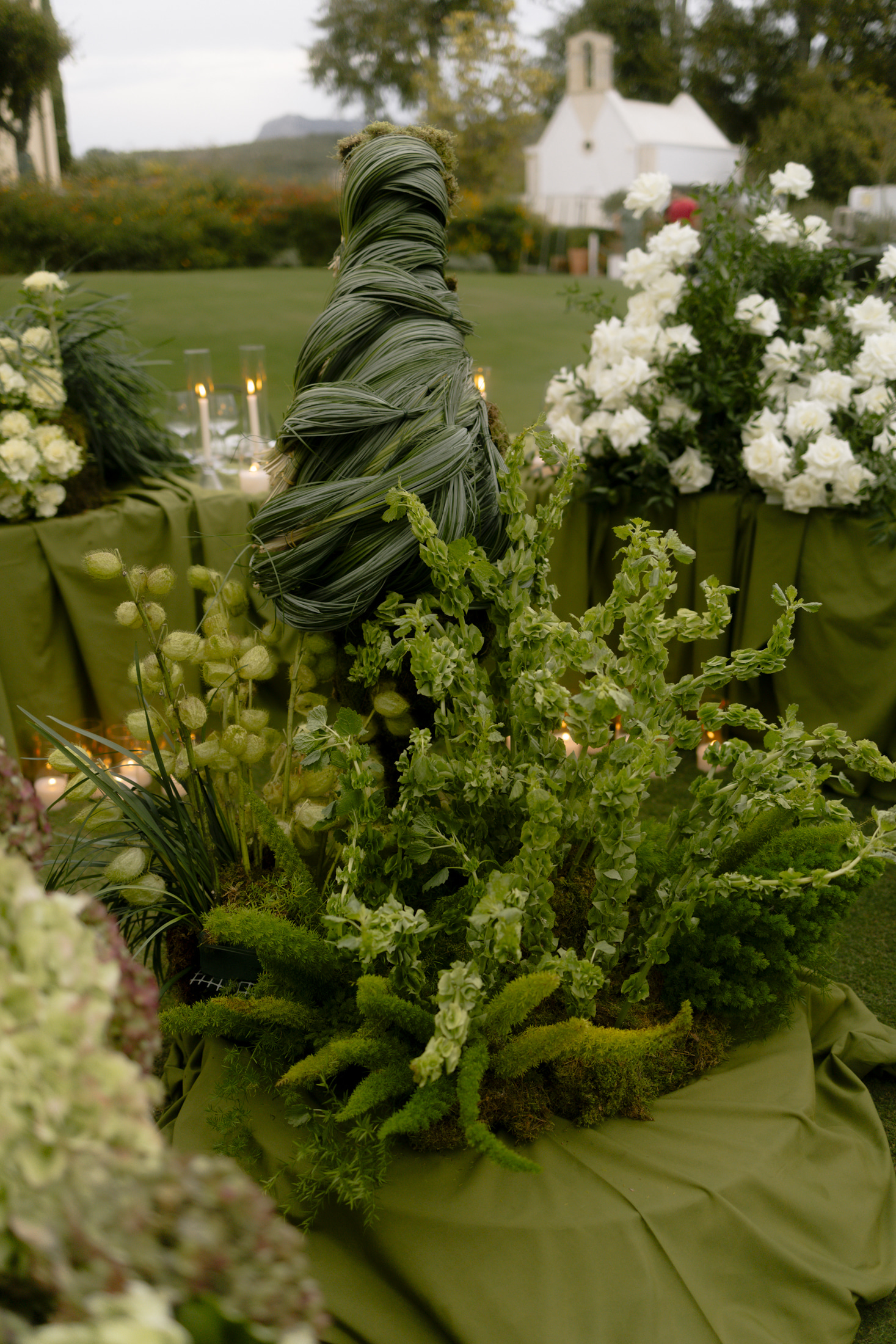 A tall, spiralling column of woven green leaves, bound with a dusting of moss, stands at the centre of a wedding table at Roxani Estate in Greece. Around its base sit sprays of bells of Ireland and papery lunaria seed pods in pale green, while on the right a cluster of white garden roses spills over a olive-green linen-draped table. Softly lit candles glow in glass hurricanes beyond, with a white chapel and a sweep of lawn blurred in the evening light.
