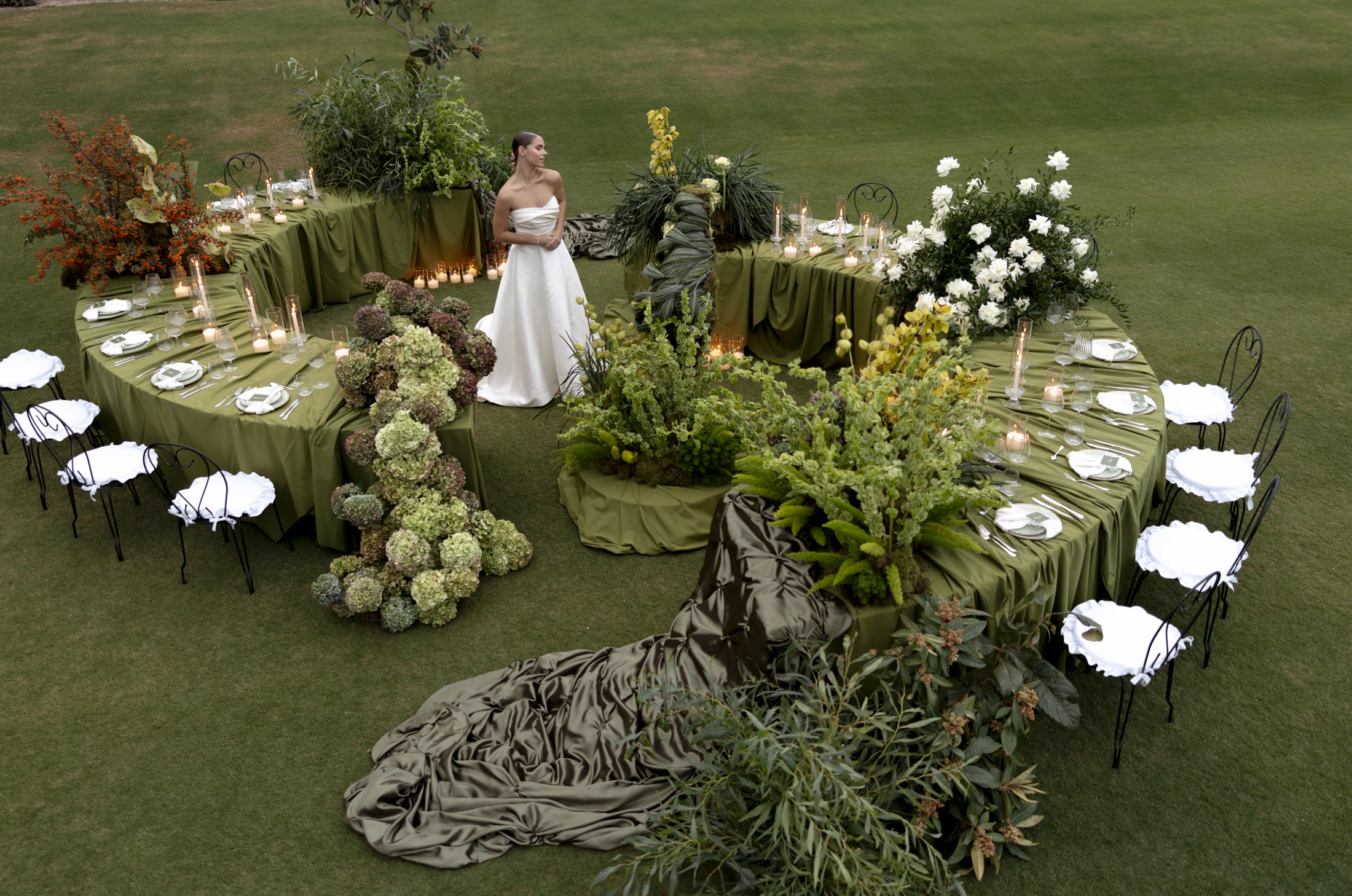 An elevated view across a lawn at Roxani Estate in Greece, where a bride in a white strapless satin gown stands among sculptural wedding tables draped in olive-green cloth. Clusters of green and burgundy hydrangeas, white roses, tall ferns and silvery eucalyptus form lush, layered centrepieces, with candles glowing along the tables and a long dark-olive satin train spilling across the grass. White ruffled-seat chairs complete the lush, tonal wedding setting.