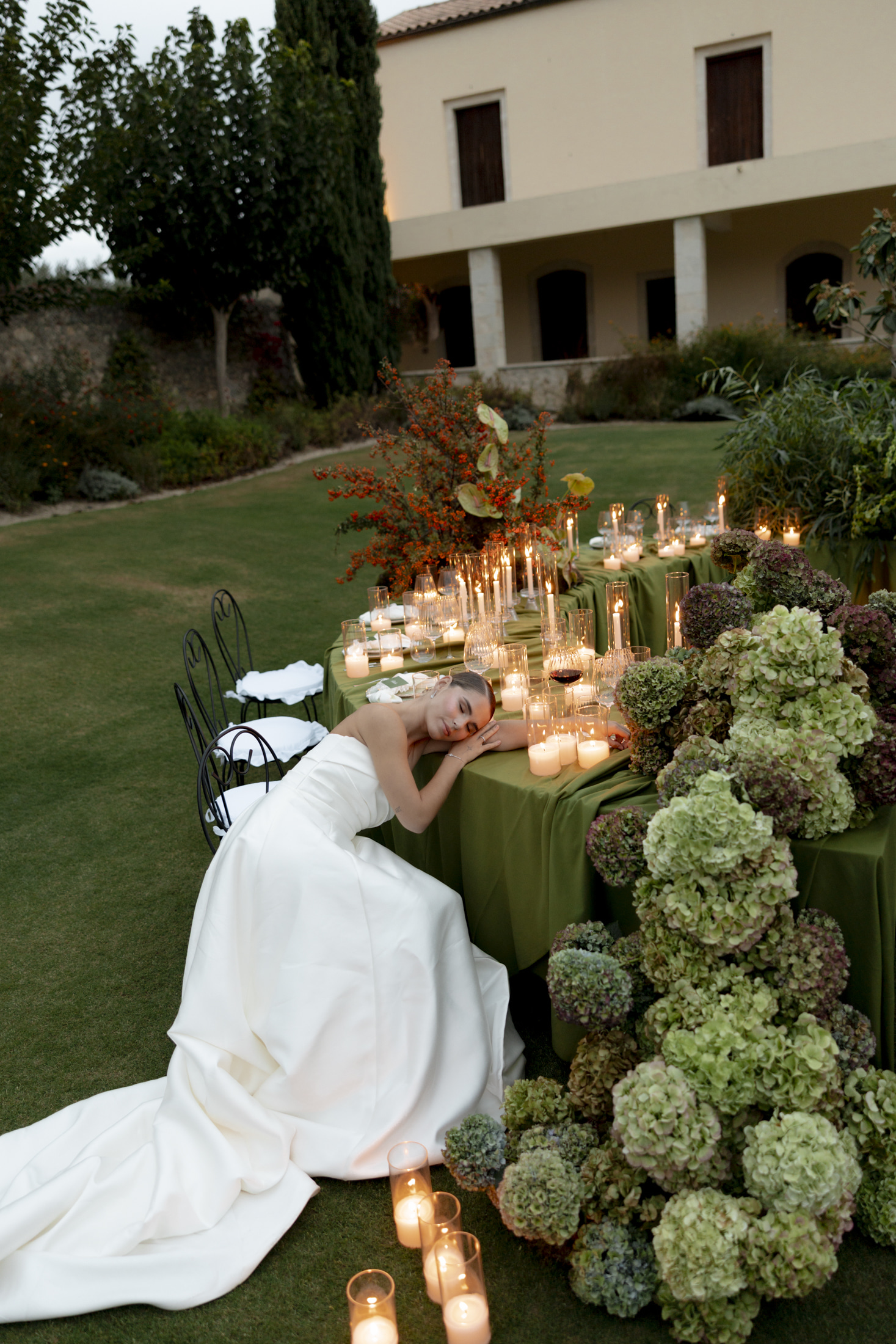 A bride in a strapless white satin gown rests her head on her arm at a long olive-green draped table on a lawn at Roxani Estate in Greece, one of the wedding tables in the Green Edit styling. Along the table, tall pillar candles and glass hurricane lanterns glow among wine glasses, and at the far end a cascade of green and plum-tinged hydrangeas spills across the cloth. A sprawling branch of red berries and dark-veined leaves rises behind, with an evening garden softly blurred beyond.