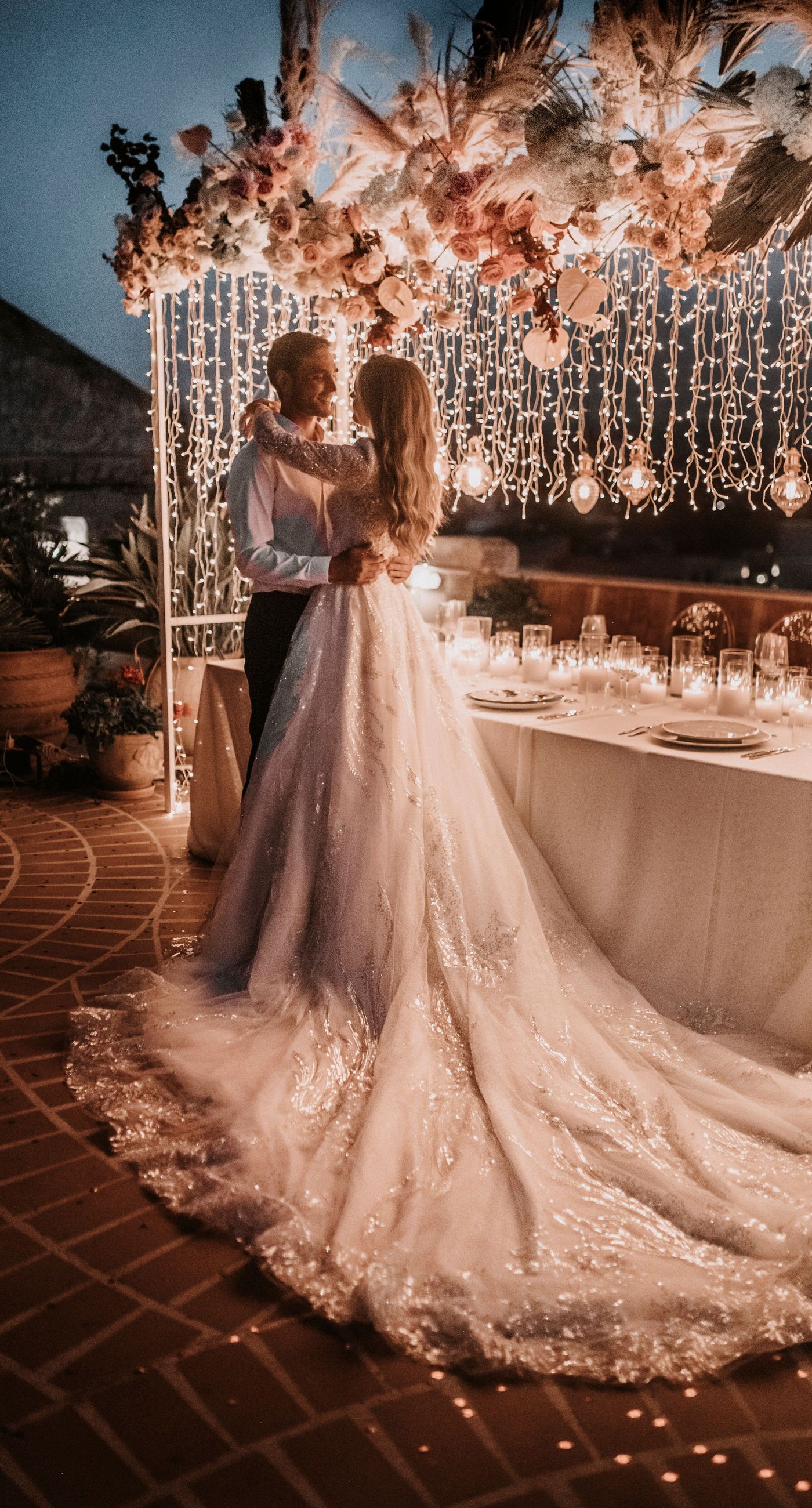 Agata & Paul - In a breathtaking celebration of love and nature's artistry, this exquisite floral masterpiece captures the essence of romance amidst a serene seaside backdrop. A delicate arch of blush pink and ivory blooms, interwoven with deep burgundy accents, frames the joyous moment, creating a harmonious blend of elegance and exuberance. The petals flutter like confetti through the air, adding a whimsical touch to the setting's charm. The terracotta and ivory-tiled floor provides a grounding contrast, enhancing the ethereal ambiance. It's a testament to the beauty that can be achieved when blossoms and emotions are artfully intertwined, making for a captivating introduction to a florist's portfolio. - Photo 6
