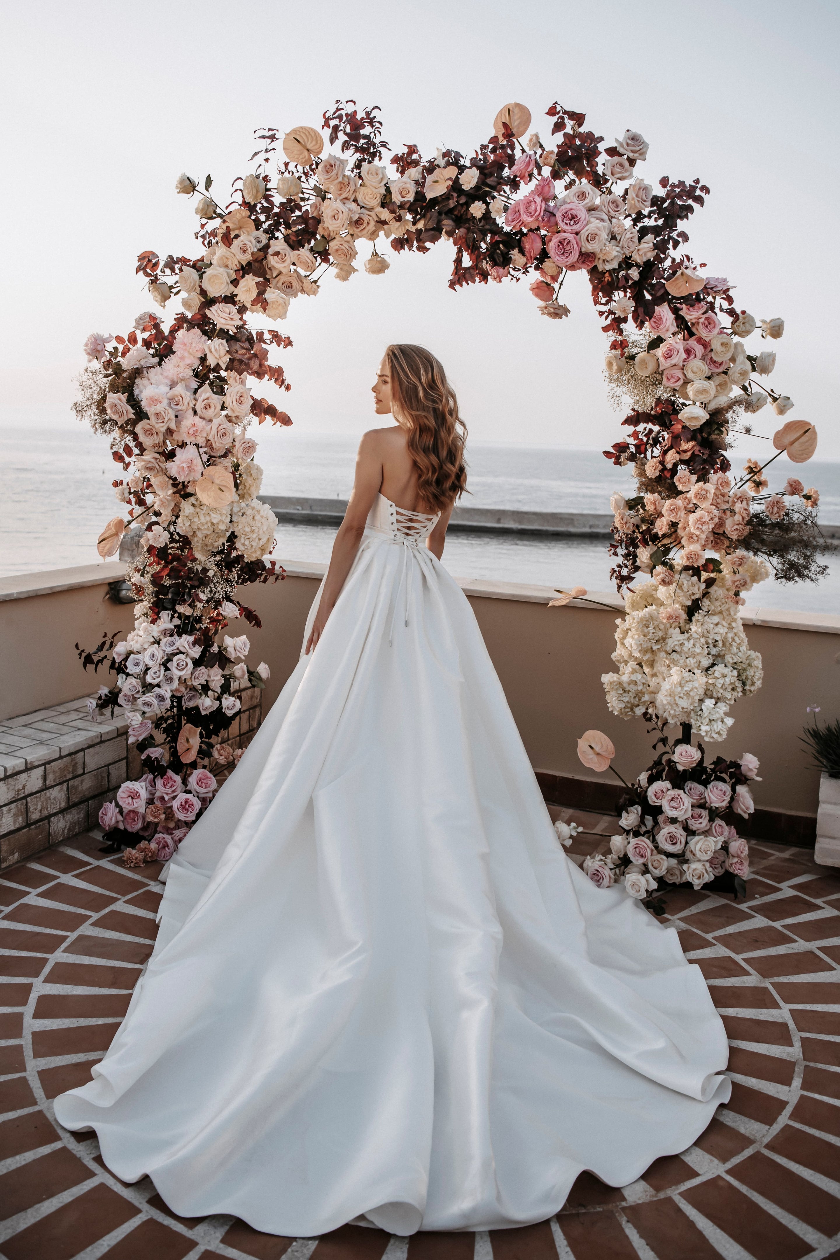 A bride in a strapless satin ball gown with a corseted lace-up back stands on a terrace with her long wavy hair falling over one shoulder, gazing out toward the sea. She faces a large round arch covered in cream and blush garden roses, white hydrangeas, anthurium and deep burgundy foliage, with a bridal arch at Pandora Suites in Greece, overlooking the calm water.