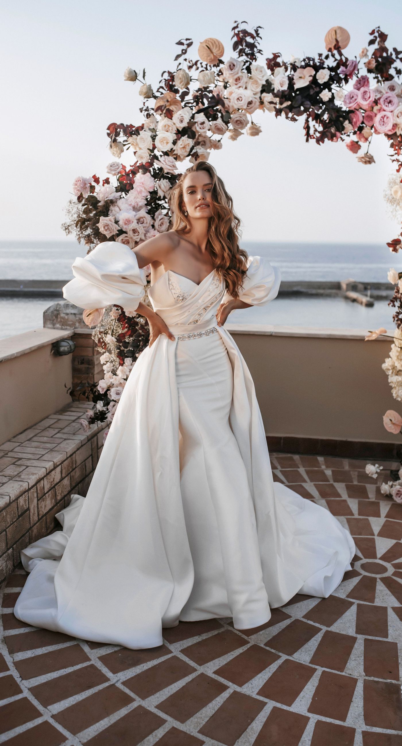 Agata & Paul - In a breathtaking celebration of love and nature's artistry, this exquisite floral masterpiece captures the essence of romance amidst a serene seaside backdrop. A delicate arch of blush pink and ivory blooms, interwoven with deep burgundy accents, frames the joyous moment, creating a harmonious blend of elegance and exuberance. The petals flutter like confetti through the air, adding a whimsical touch to the setting's charm. The terracotta and ivory-tiled floor provides a grounding contrast, enhancing the ethereal ambiance. It's a testament to the beauty that can be achieved when blossoms and emotions are artfully intertwined, making for a captivating introduction to a florist's portfolio. - Photo 2