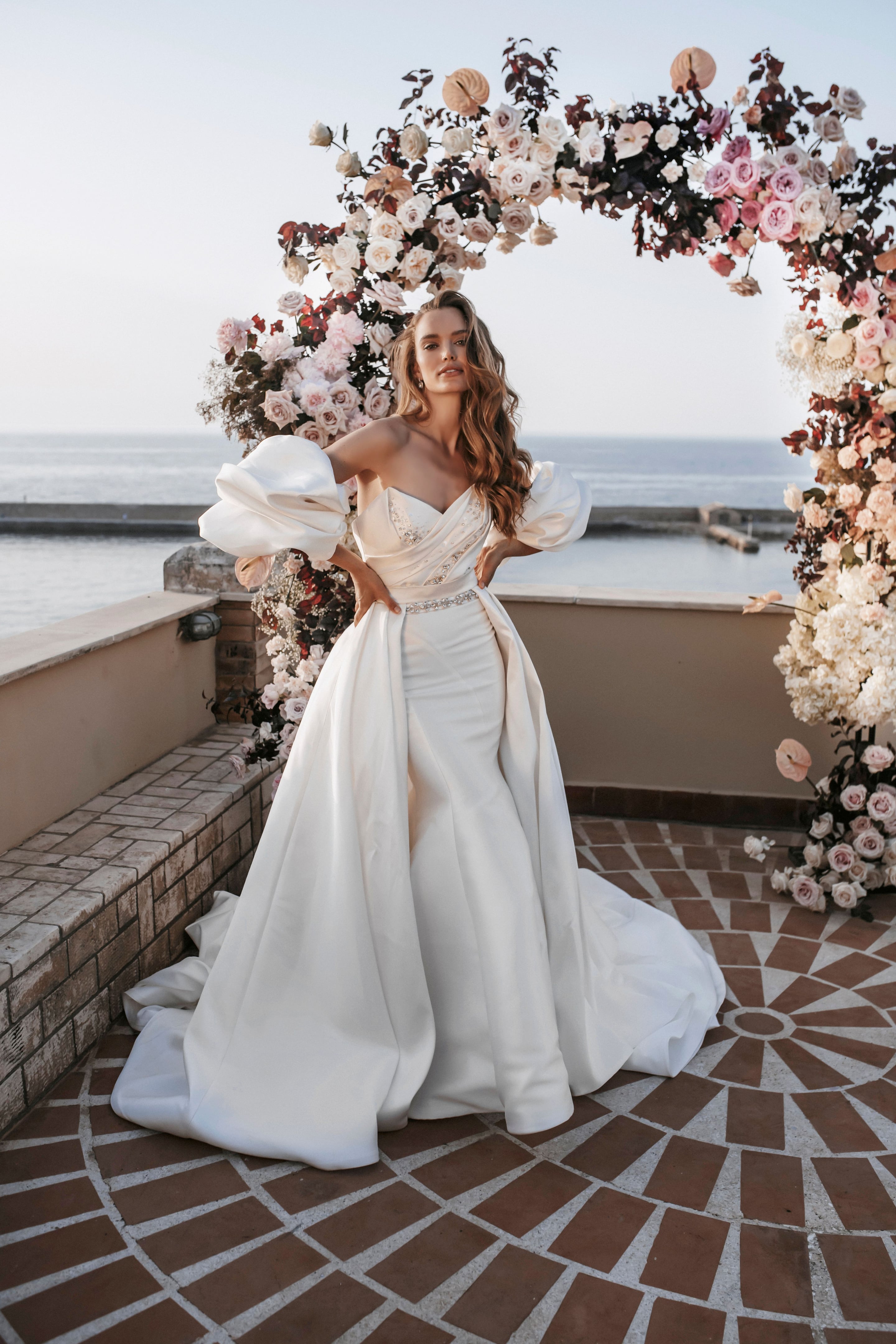 A bride with long, loosely waved honey-blonde hair stands with her hands on her hips in a satin strapless gown with puffed sleeves and a jewelled waistband, in front of a curved arch of blush, cream and deep burgundy roses and hydrangeas. The wedding flowers frame a terrace at Pandora Suites in Greece, with calm sea and a pale sky behind her.