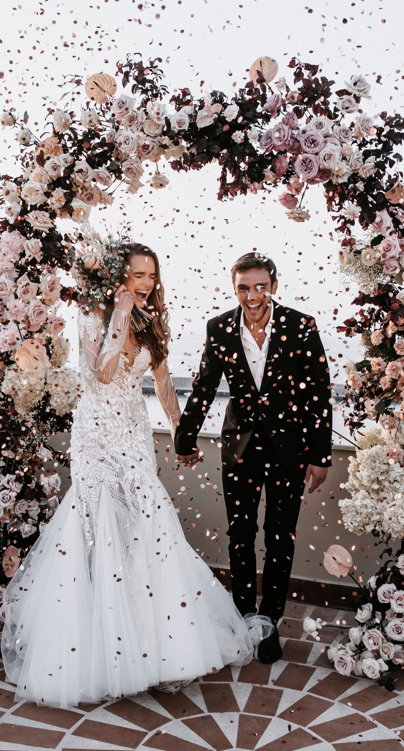 Agata & Paul - In a breathtaking celebration of love and nature's artistry, this exquisite floral masterpiece captures the essence of romance amidst a serene seaside backdrop. A delicate arch of blush pink and ivory blooms, interwoven with deep burgundy accents, frames the joyous moment, creating a harmonious blend of elegance and exuberance. The petals flutter like confetti through the air, adding a whimsical touch to the setting's charm. The terracotta and ivory-tiled floor provides a grounding contrast, enhancing the ethereal ambiance. It's a testament to the beauty that can be achieved when blossoms and emotions are artfully intertwined, making for a captivating introduction to a florist's portfolio. - Photo 4