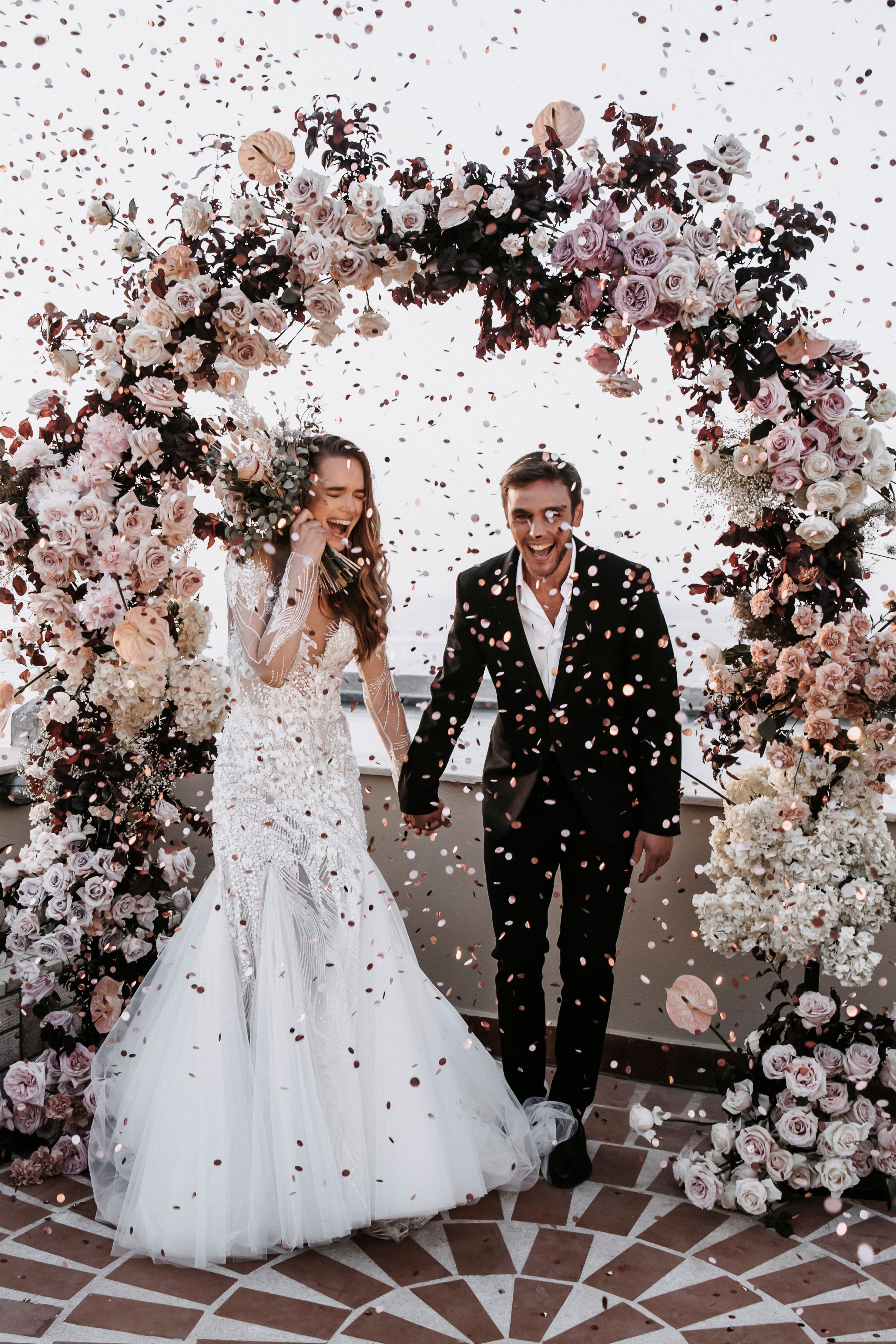 A bride and groom laugh together as pink and gold confetti falls around them, holding hands beneath a tall arch of dusky pink garden roses, cream hydrangeas and deep burgundy foliage. The bride, in a long-sleeved, heavily beaded white gown, holds a bouquet of eucalyptus and pale blooms against her face. This wedding arch and bridal bouquet are from Pandora Suites in Greece.