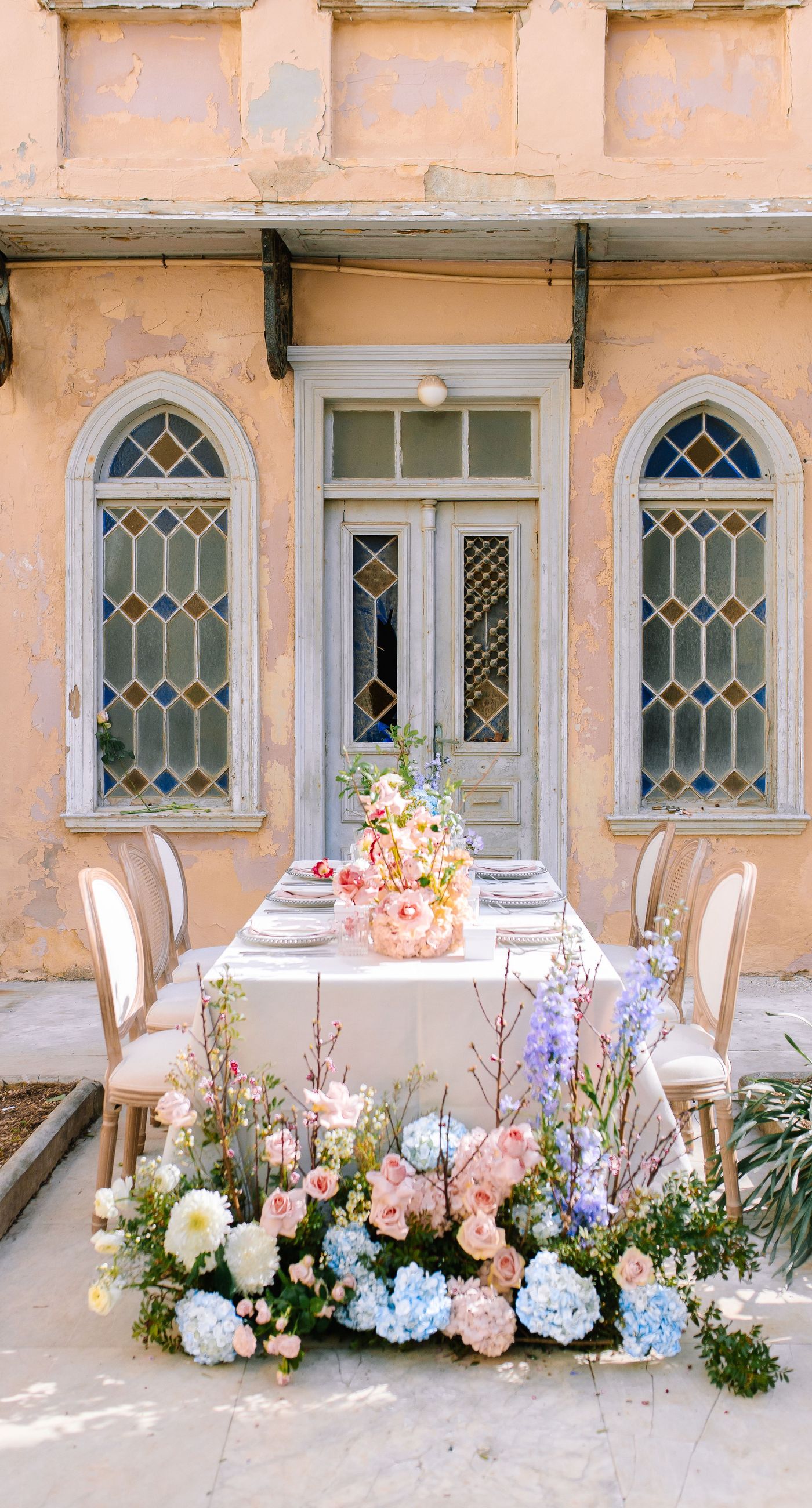 Alysia & Fraunk - Amidst the backdrop of a weathered, pale peach facade with antique windows, an enchanting scene unfolds. The romantic embrace of a couple is tenderly framed by a symphony of pastel-hued florals—soft pinks, gentle blues, and creamy whites—exuding timeless elegance. The delicate wild arrangements accentuate the ethereal beauty of the bridal dress, while the muted tones of the aged architecture blend seamlessly with the lush, garden-like display. This harmonious composition captures the essence of rustic romance, showcasing the florist's ability to weave nature's hues into a visual poem of love. - Photo 7