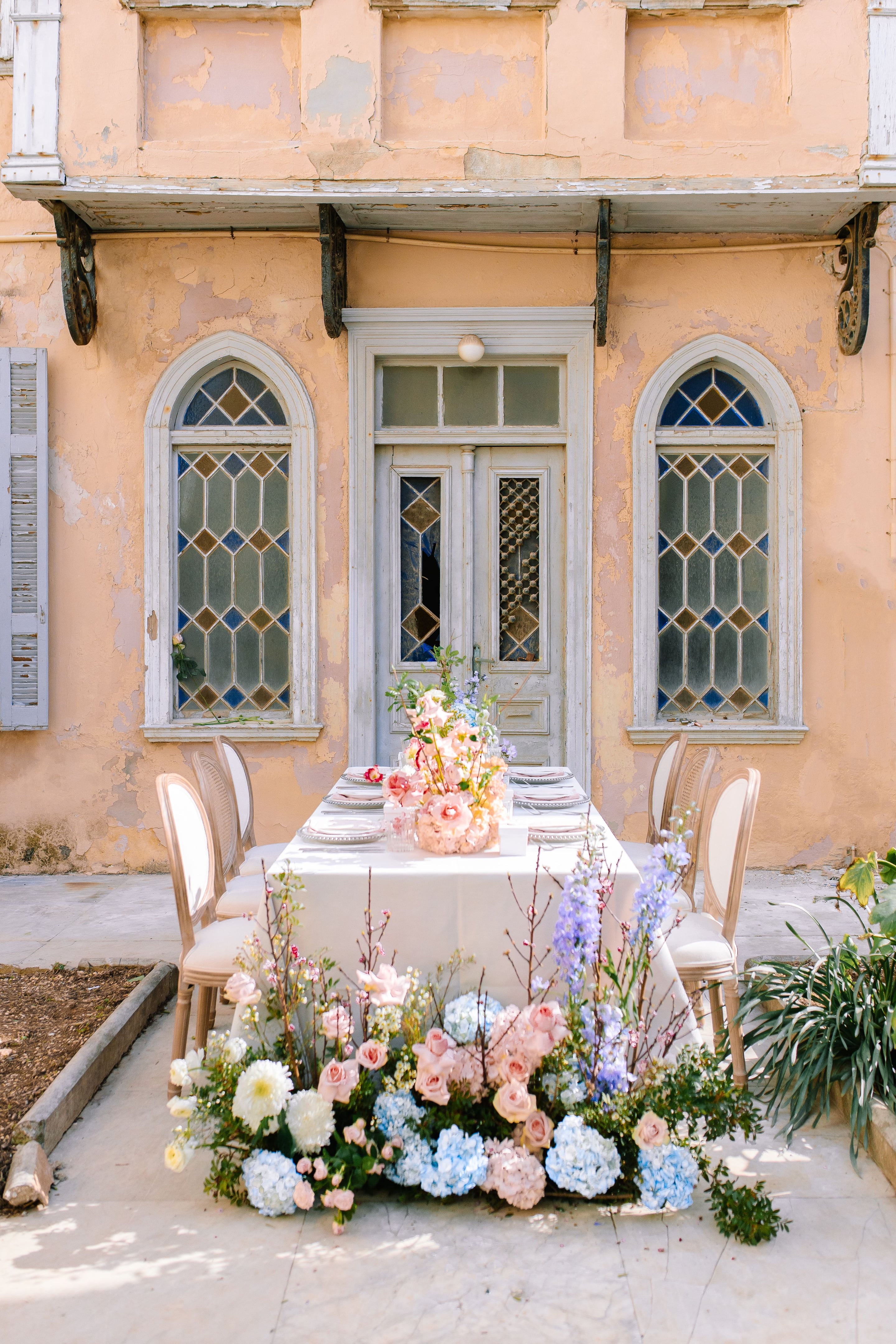 A long table dressed in white linen on a sunlit stone terrace in Greece, set with wedding table flowers in soft pastels. Along the table's base runs a low, lush arrangement of pale pink garden roses, white dahlias, blue hydrangeas, and lavender-blue delphinium, with bare peach branches and greenery rising through it. Behind, a peach-washed wall with peeling plaster frames a tall wooden door and leaded stained-glass windows, and cane-backed chairs line the table.