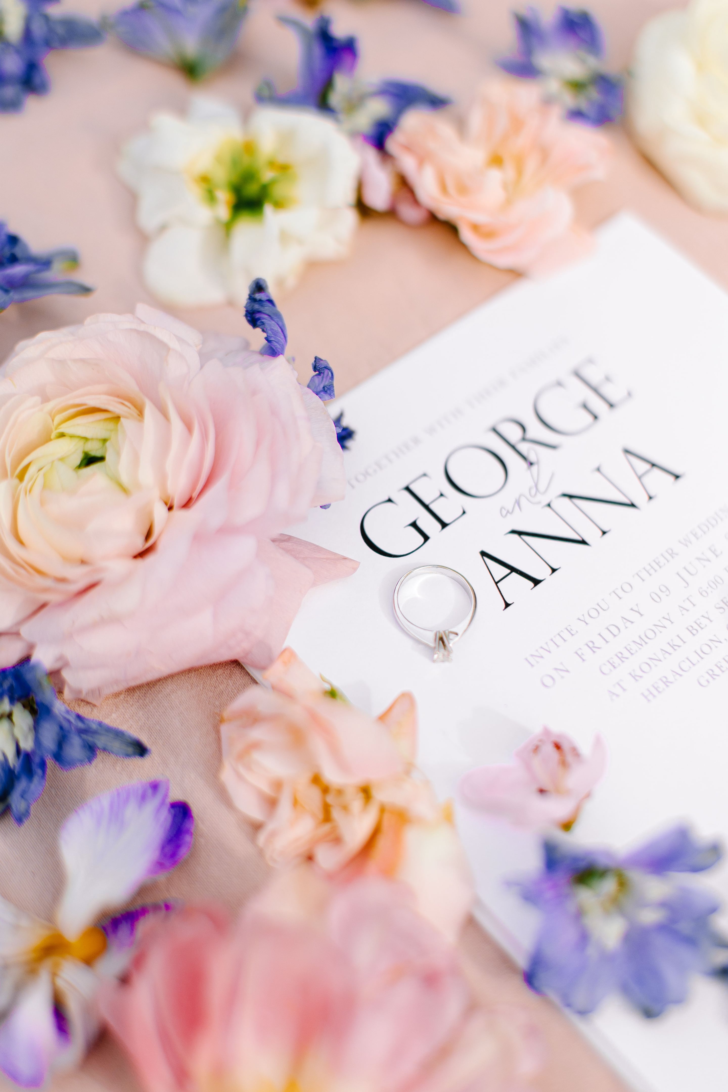 A flat-lay of soft pink ranunculus, cream blooms and small purple delphinium spires resting on a blush-coloured cloth, with a silver ring placed on a white wedding invitation. The invitation's bold black lettering reads "George and Anna" with smaller text beneath it, and the whole scene is softly lit. These are wedding flowers in Greece, styled alongside the couple's stationery in Heraclion, Crete.
