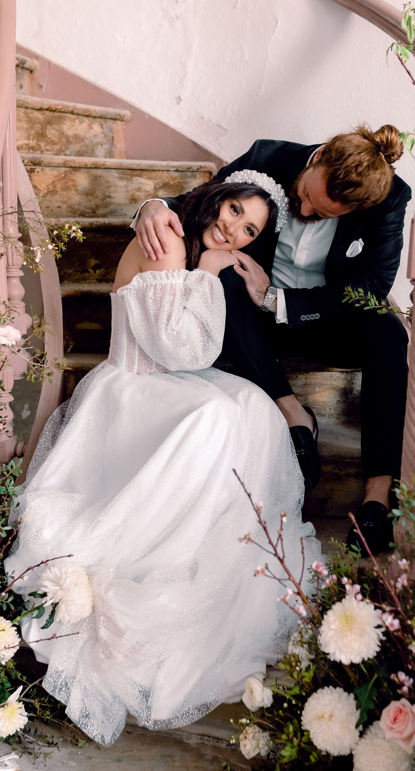 Alysia & Fraunk - Amidst the backdrop of a weathered, pale peach facade with antique windows, an enchanting scene unfolds. The romantic embrace of a couple is tenderly framed by a symphony of pastel-hued florals—soft pinks, gentle blues, and creamy whites—exuding timeless elegance. The delicate wild arrangements accentuate the ethereal beauty of the bridal dress, while the muted tones of the aged architecture blend seamlessly with the lush, garden-like display. This harmonious composition captures the essence of rustic romance, showcasing the florist's ability to weave nature's hues into a visual poem of love. - Photo 10