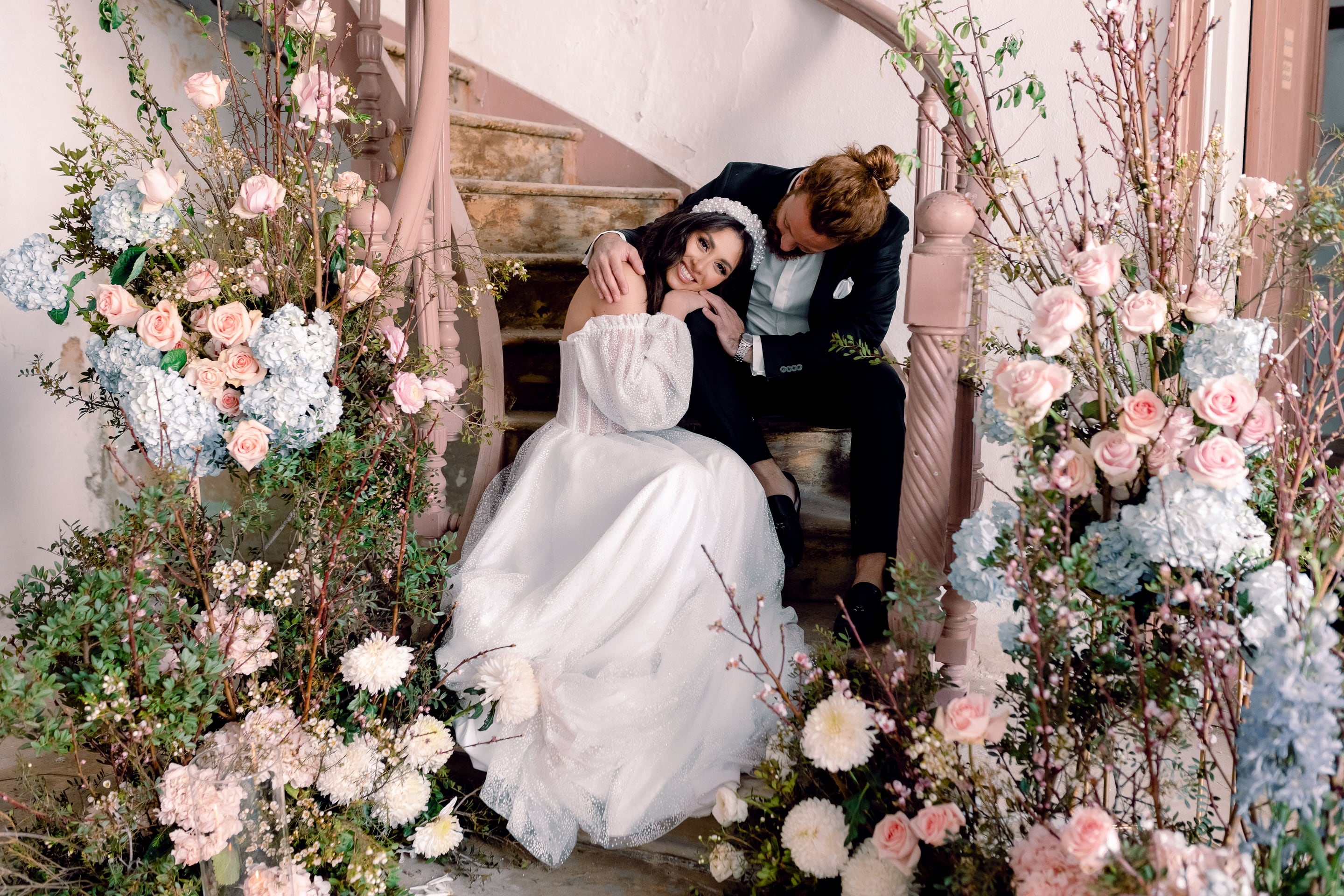 A bride in a sparkly white gown with puffed sheer sleeves and a pearl headband rests her cheek against the shoulder of a groom in a dark suit, who leans in beside her on a weathered, rose-painted spiral staircase. Both are smiling softly. The staircase is framed on each side by lush arrangements of pale pink garden roses, blush hydrangeas, white chrysanthemums, and flowering branches, all part of the wedding flower styling in Greece.