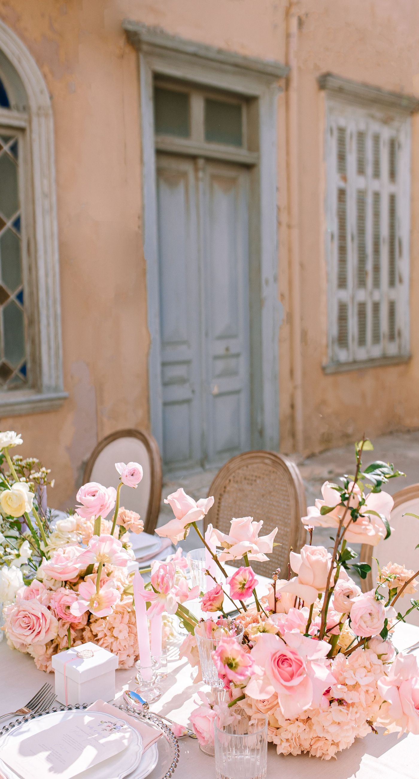 Alysia & Fraunk - Amidst the backdrop of a weathered, pale peach facade with antique windows, an enchanting scene unfolds. The romantic embrace of a couple is tenderly framed by a symphony of pastel-hued florals—soft pinks, gentle blues, and creamy whites—exuding timeless elegance. The delicate wild arrangements accentuate the ethereal beauty of the bridal dress, while the muted tones of the aged architecture blend seamlessly with the lush, garden-like display. This harmonious composition captures the essence of rustic romance, showcasing the florist's ability to weave nature's hues into a visual poem of love. - Photo 5