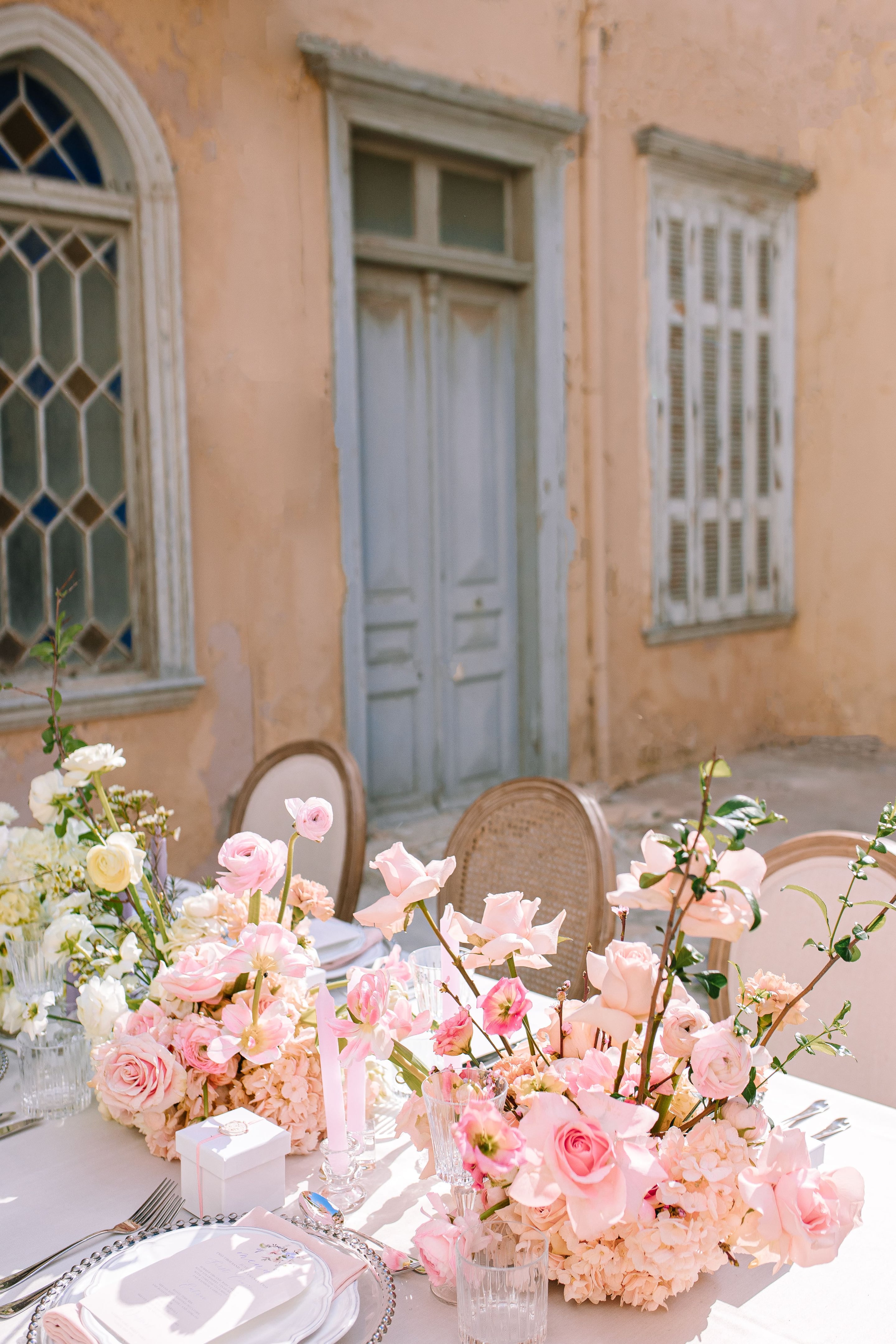 Low pink and cream centrepieces of garden roses, ranunculus, peonies and peach hydrangeas fill a long wedding table set outdoors in Greece, with tall branches of green foliage rising through the arrangements. Pale pink taper candles stand in glass holders beside a small white gift box tied with a pink ribbon, and clear glasses and silver cutlery sit on a soft linen cloth. Behind the table, a weathered ochre wall, a pale blue wooden door and a shuttered window catch warm, gentle sunlight, giving the scene a soft, romantic mood.
