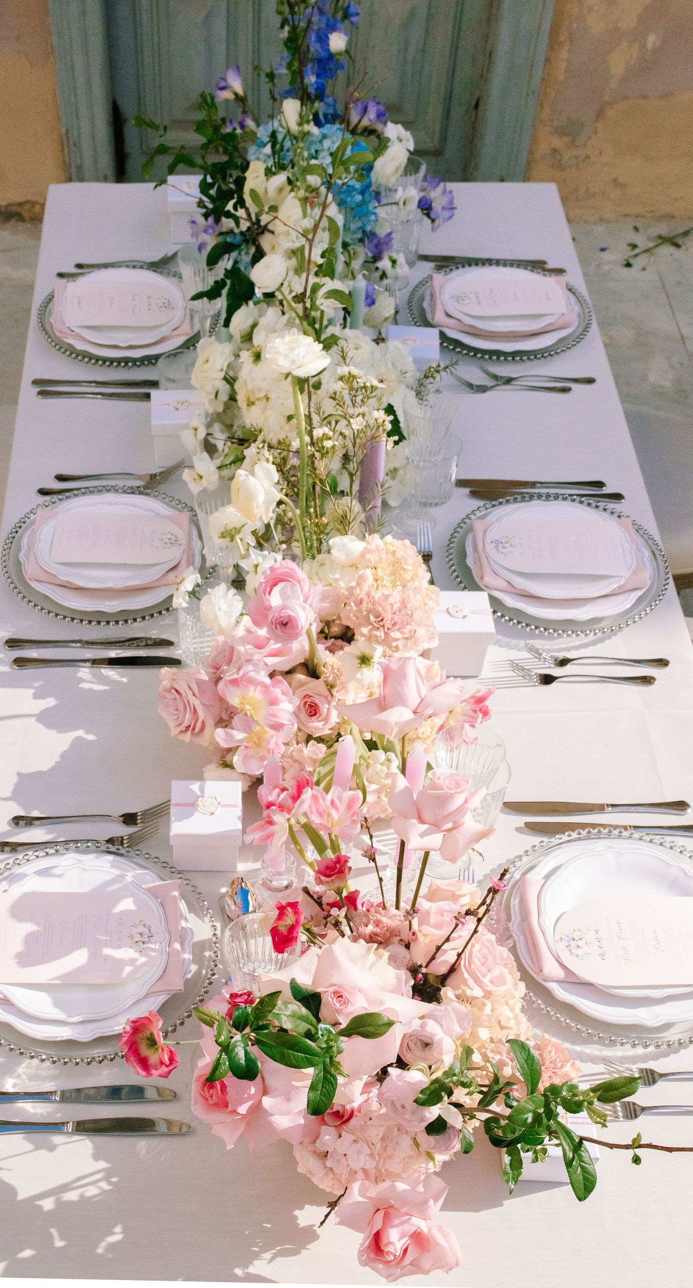 Alysia & Fraunk - Amidst the backdrop of a weathered, pale peach facade with antique windows, an enchanting scene unfolds. The romantic embrace of a couple is tenderly framed by a symphony of pastel-hued florals—soft pinks, gentle blues, and creamy whites—exuding timeless elegance. The delicate wild arrangements accentuate the ethereal beauty of the bridal dress, while the muted tones of the aged architecture blend seamlessly with the lush, garden-like display. This harmonious composition captures the essence of rustic romance, showcasing the florist's ability to weave nature's hues into a visual poem of love. - Photo 8