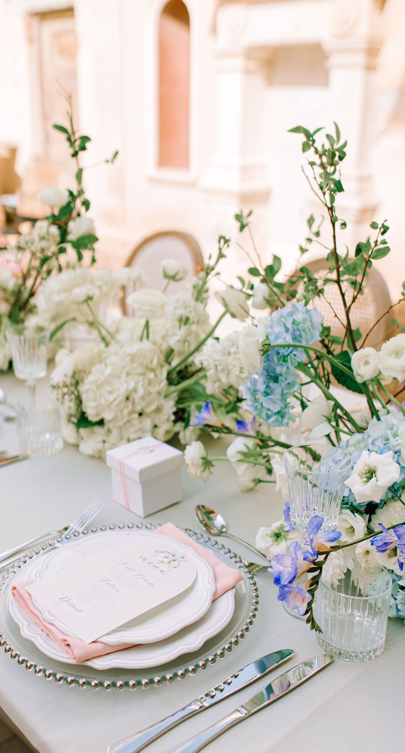 Alysia & Fraunk - Amidst the backdrop of a weathered, pale peach facade with antique windows, an enchanting scene unfolds. The romantic embrace of a couple is tenderly framed by a symphony of pastel-hued florals—soft pinks, gentle blues, and creamy whites—exuding timeless elegance. The delicate wild arrangements accentuate the ethereal beauty of the bridal dress, while the muted tones of the aged architecture blend seamlessly with the lush, garden-like display. This harmonious composition captures the essence of rustic romance, showcasing the florist's ability to weave nature's hues into a visual poem of love. - Photo 4