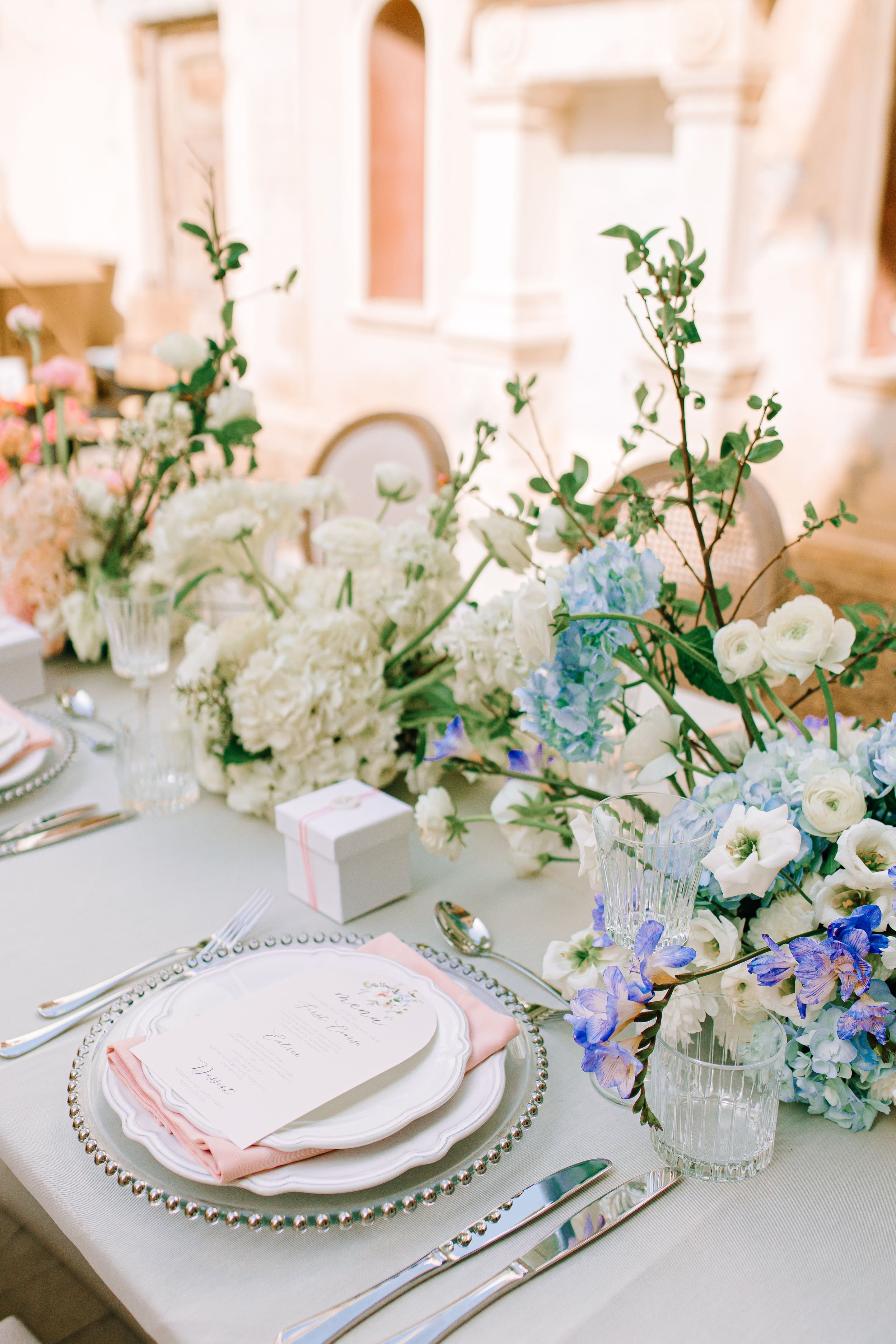 Wedding table flowers in Greece: a tall, airy centrepiece of white hydrangeas, white ranunculus and blue hydrangeas, with spires of purple freesia and bare green branches rising through them. A white favour box tied with a pale pink ribbon sits on the linen tablecloth beside a place setting with a pink napkin, a beaded glass charger and a printed menu card. The soft, sunlit background and pale blue and cream tones give the table a calm, romantic feel.
