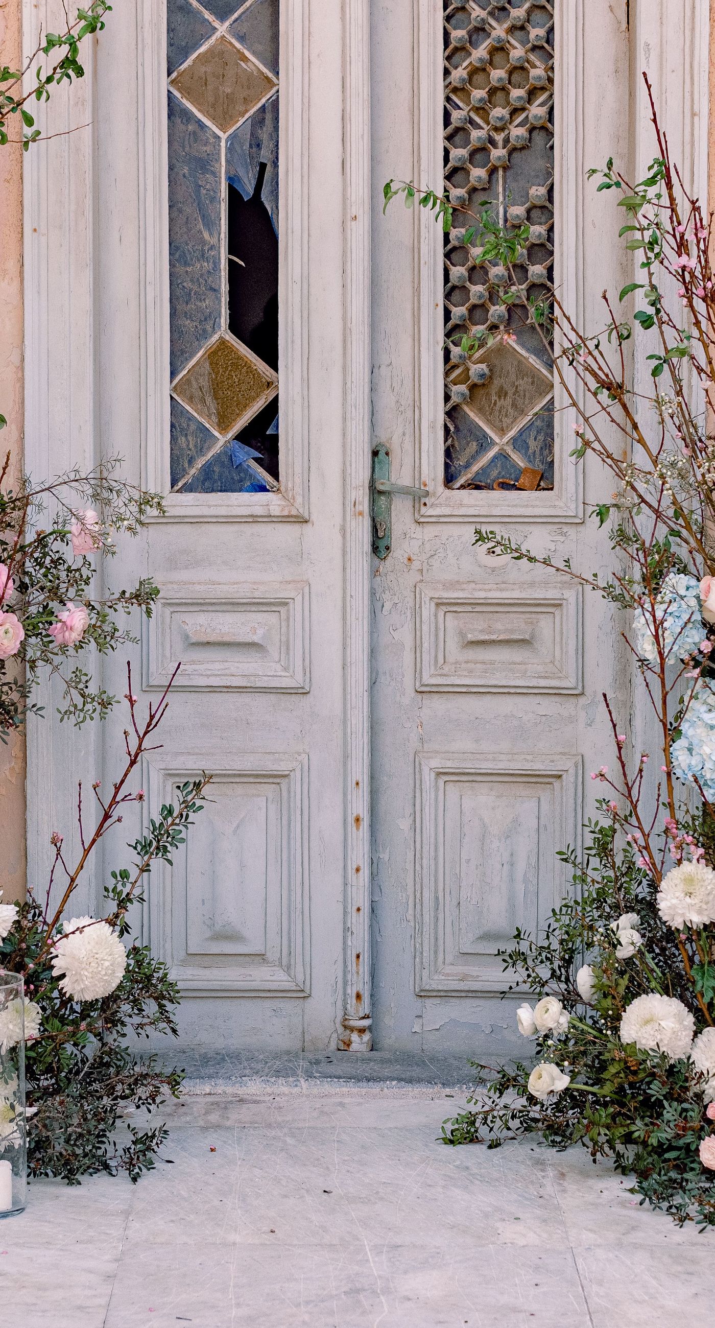 Alysia & Fraunk - Amidst the backdrop of a weathered, pale peach facade with antique windows, an enchanting scene unfolds. The romantic embrace of a couple is tenderly framed by a symphony of pastel-hued florals—soft pinks, gentle blues, and creamy whites—exuding timeless elegance. The delicate wild arrangements accentuate the ethereal beauty of the bridal dress, while the muted tones of the aged architecture blend seamlessly with the lush, garden-like display. This harmonious composition captures the essence of rustic romance, showcasing the florist's ability to weave nature's hues into a visual poem of love. - Photo 2