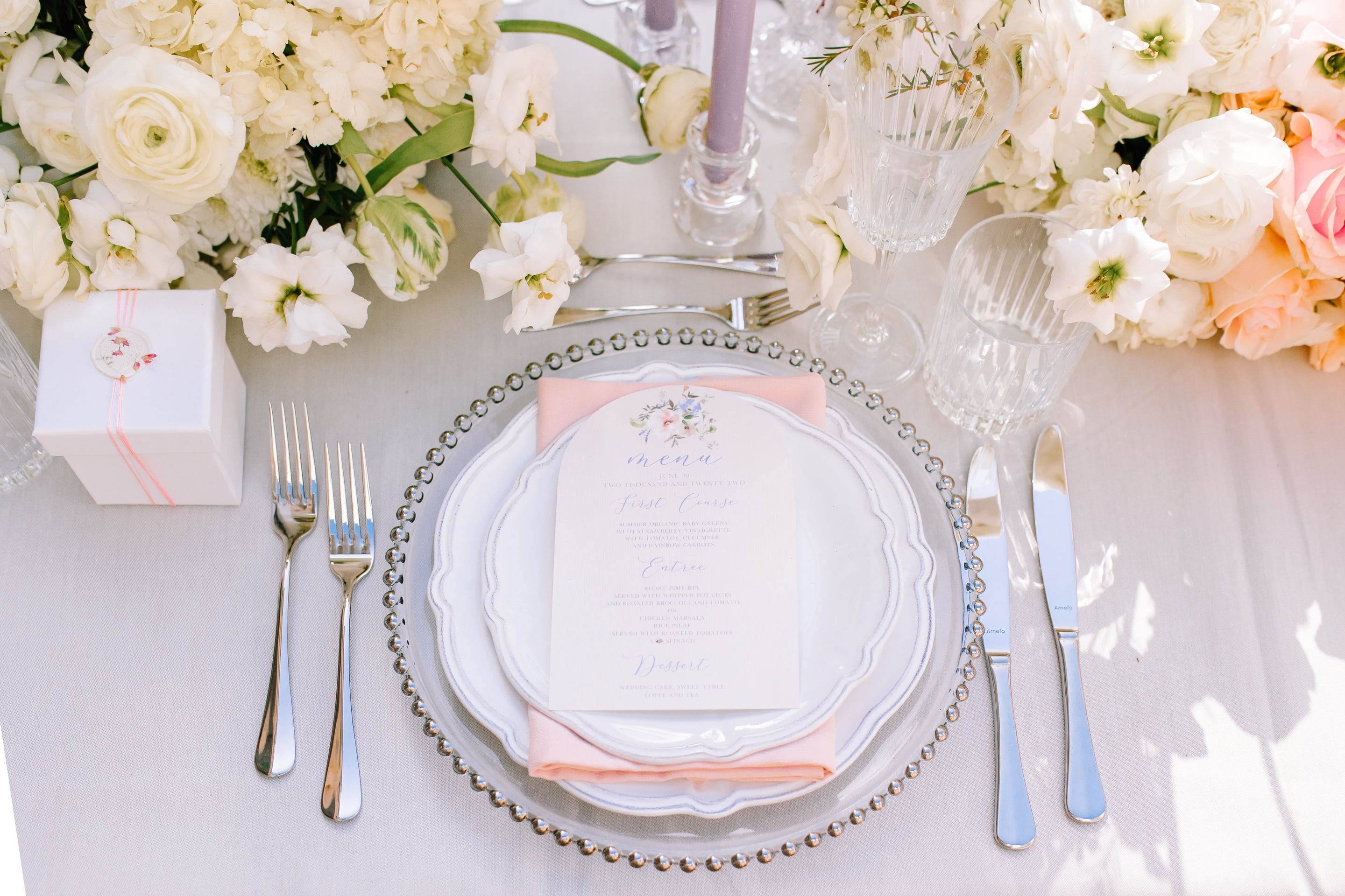 A wedding table setting in Greece, dressed in soft whites and blush: a place setting with a beaded glass charger, layered white scalloped plates, and a blush pink napkin topped by a menu card with a small watercolour floral motif. Above it, a low centrepiece of white hydrangeas, ranunculus and lisianthus spills across the linen, beside a lilac taper candle and a small white gift box tied with pink thread.