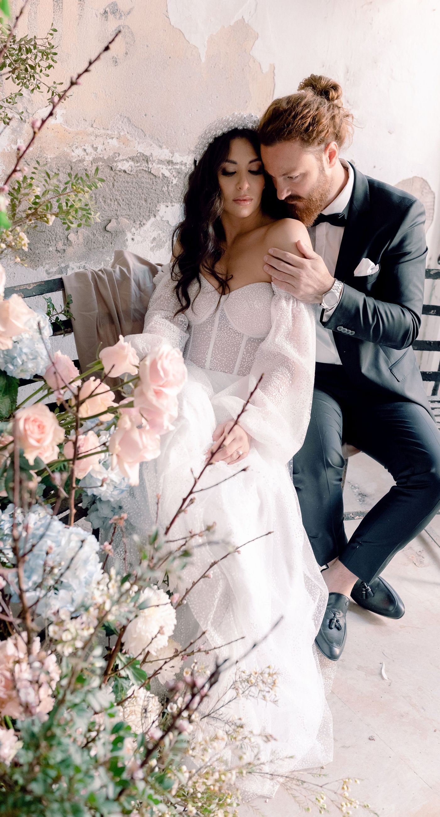 Alysia & Fraunk - Amidst the backdrop of a weathered, pale peach facade with antique windows, an enchanting scene unfolds. The romantic embrace of a couple is tenderly framed by a symphony of pastel-hued florals—soft pinks, gentle blues, and creamy whites—exuding timeless elegance. The delicate wild arrangements accentuate the ethereal beauty of the bridal dress, while the muted tones of the aged architecture blend seamlessly with the lush, garden-like display. This harmonious composition captures the essence of rustic romance, showcasing the florist's ability to weave nature's hues into a visual poem of love. - Photo 11
