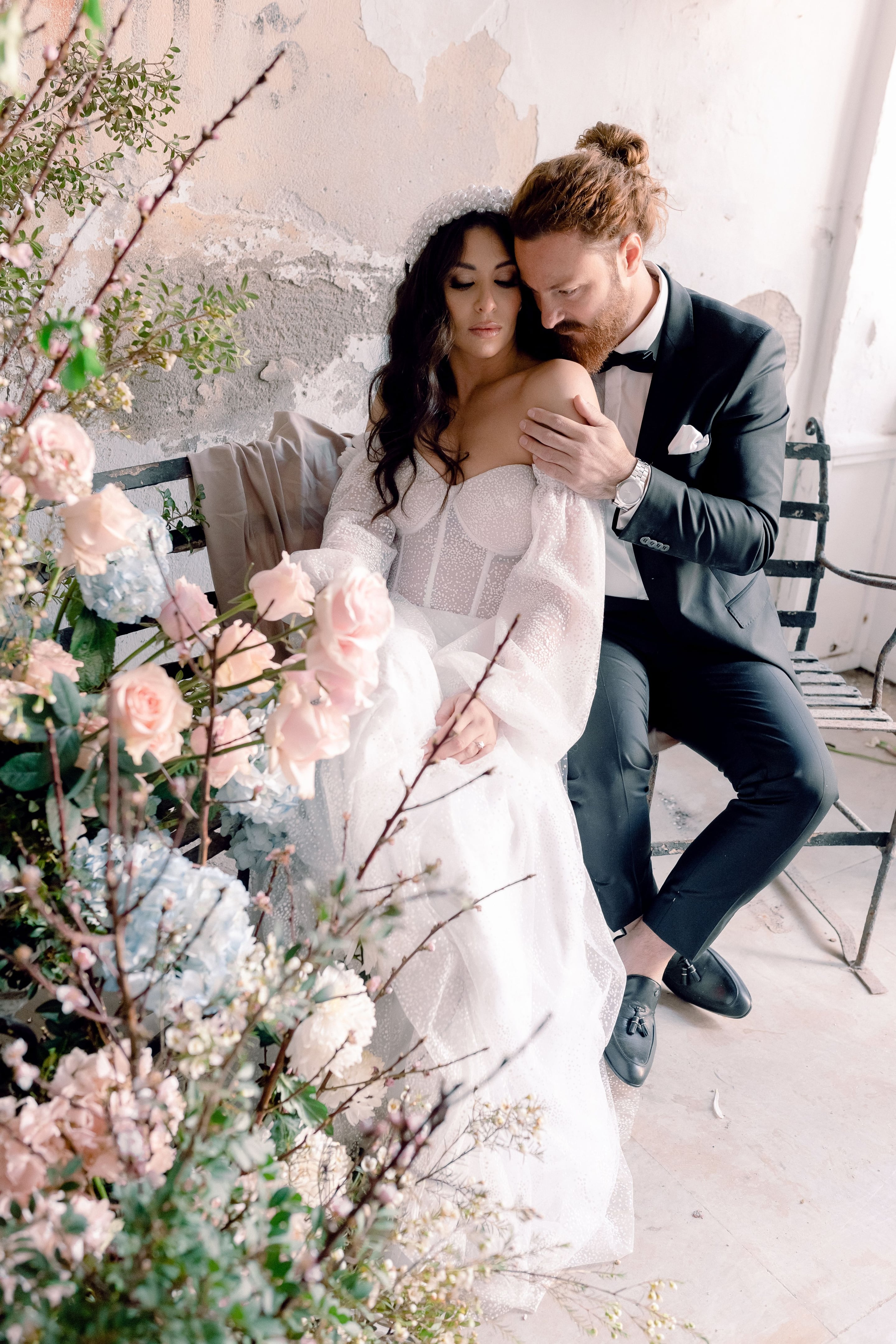 A bride in a sheer, dotted-tulle gown with puffed sleeves and a corset bodice sits on a weathered iron bench, while a groom in a dark tuxedo leans in to rest his hand on her shoulder. In the foreground, a lush arrangement of pale pink garden roses, blue hydrangeas and bare blossom branches frames the couple against a crumbling plaster wall. This wedding flower styling in Greece has a soft, airy palette of blush, pale blue and cream.