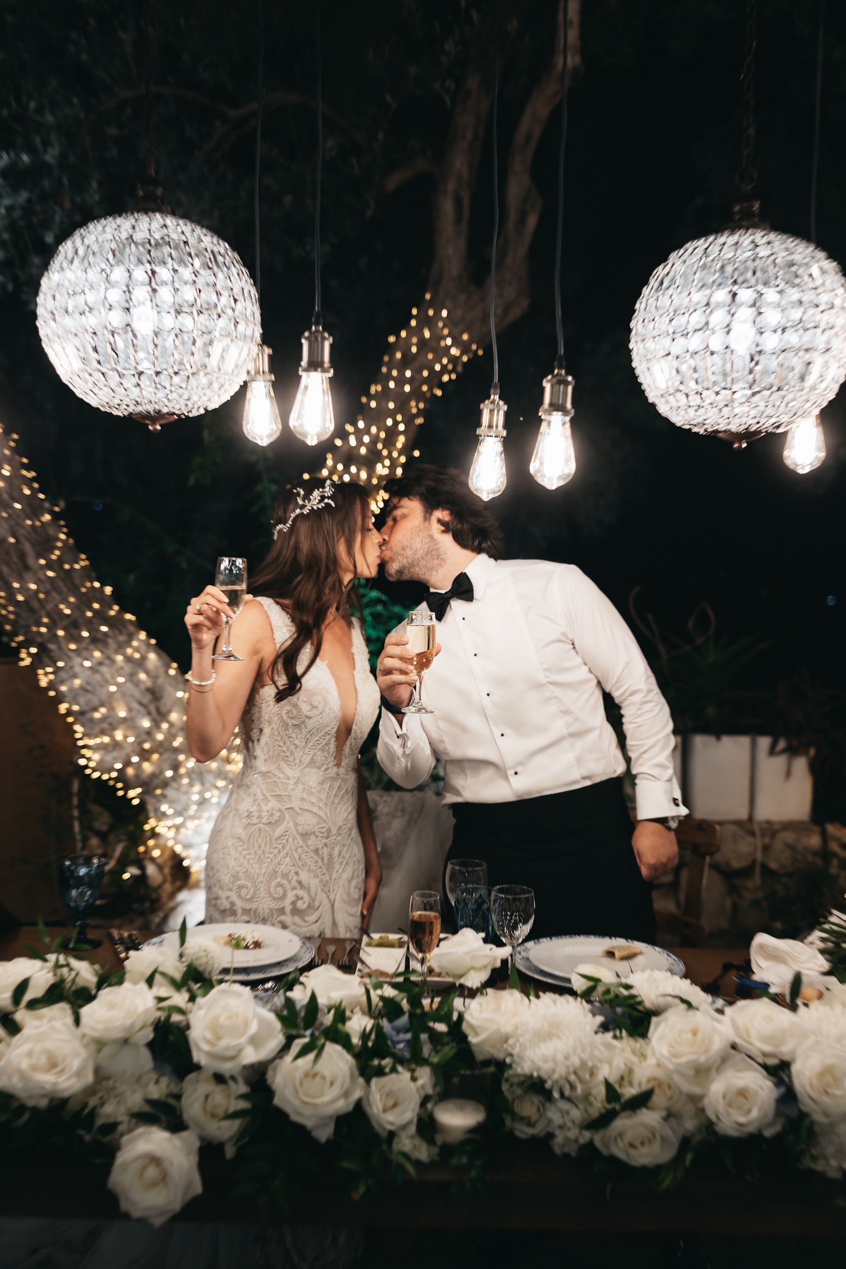 A bride and groom share a kiss at night beside a long head table at Agreco Farm in Greece, each holding a flute of champagne. Her lace gown has a deep V neckline and a delicate silver leaf headpiece, and his white shirt and black bow tie stand out against the dark sky. Above them, crystal chandeliers and bare Edison bulbs glow, and strings of fairy lights wrap the trees behind. In the foreground, a centrepiece of white garden roses, white chrysanthemums and trailing greenery sits among blue-rimmed plates and glasses, all for this wedding.