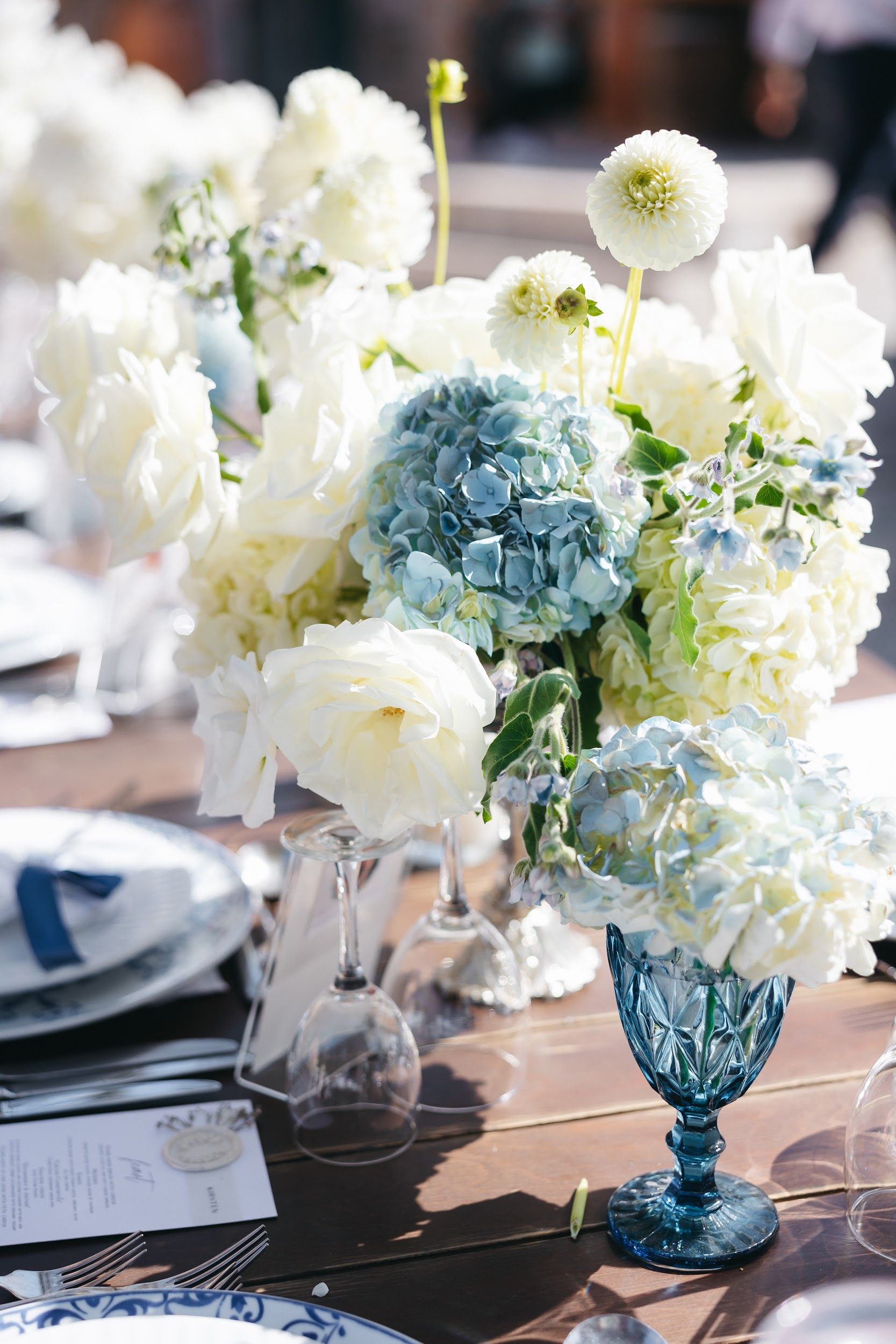 A wedding table centrepiece of creamy white garden roses and white hydrangeas, with pale blue hydrangea heads and sprigs of blue borage, rising from a faceted blue glass goblet on a rustic wooden table at Agreco Farm in Greece. Behind it, a place setting with a white plate, a navy ribbon-tied napkin and upturned clear wine glasses catches soft, bright daylight, giving the scene a calm, airy, coastal feel.