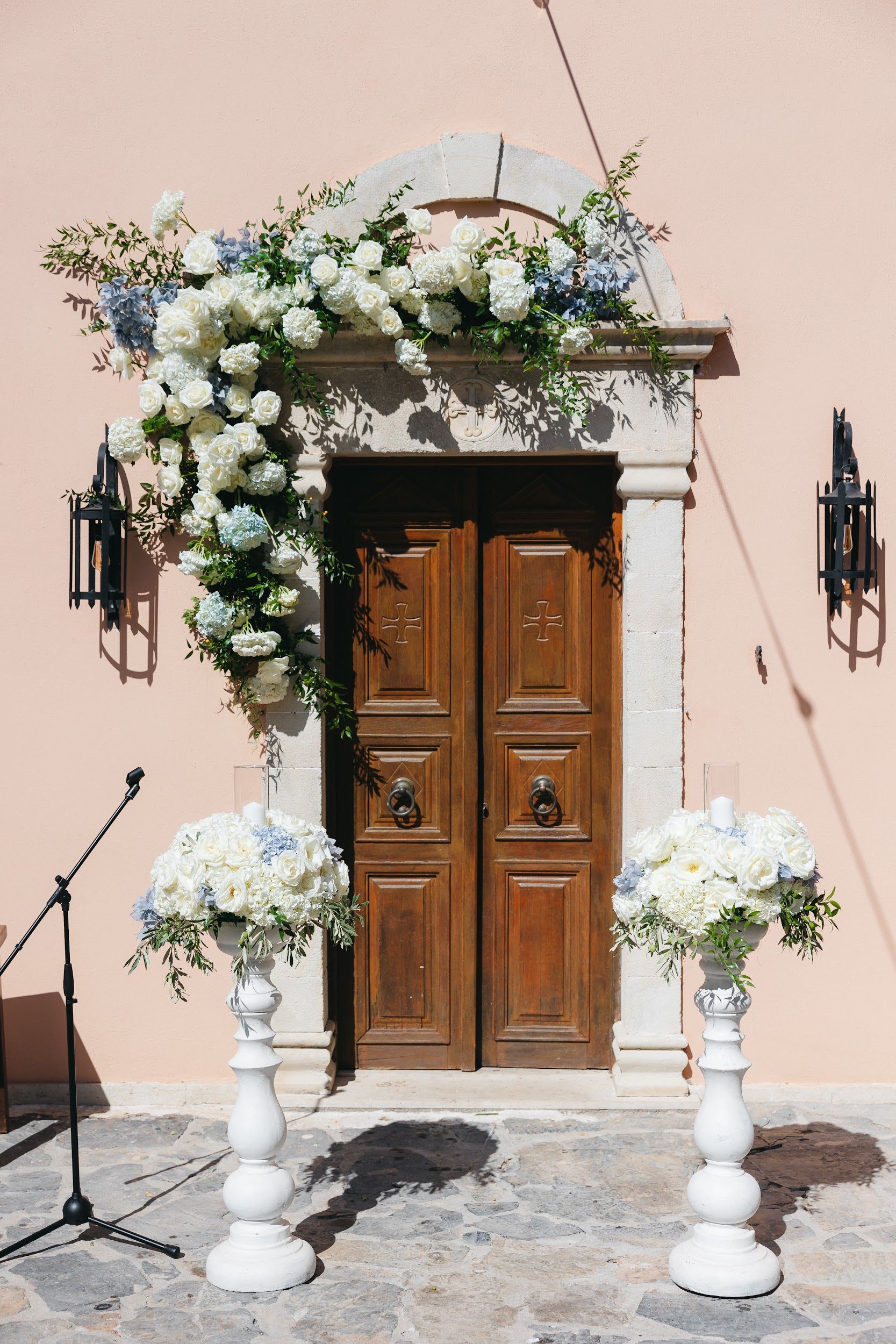 A cascading floral arch of white garden roses, white hydrangeas and soft blue hydrangeas with trailing olive branches drapes over a pale stone doorway with tall wooden double doors, the flowers spilling down the left side. Two lush white floral arrangements sit on pedestals at the foot of the doors, with a glowing candle in a glass hurricane at the centre of each. This wedding flower display, on a sunlit pink wall at Agreco Farm in Greece, has a fresh, elegant feel.
