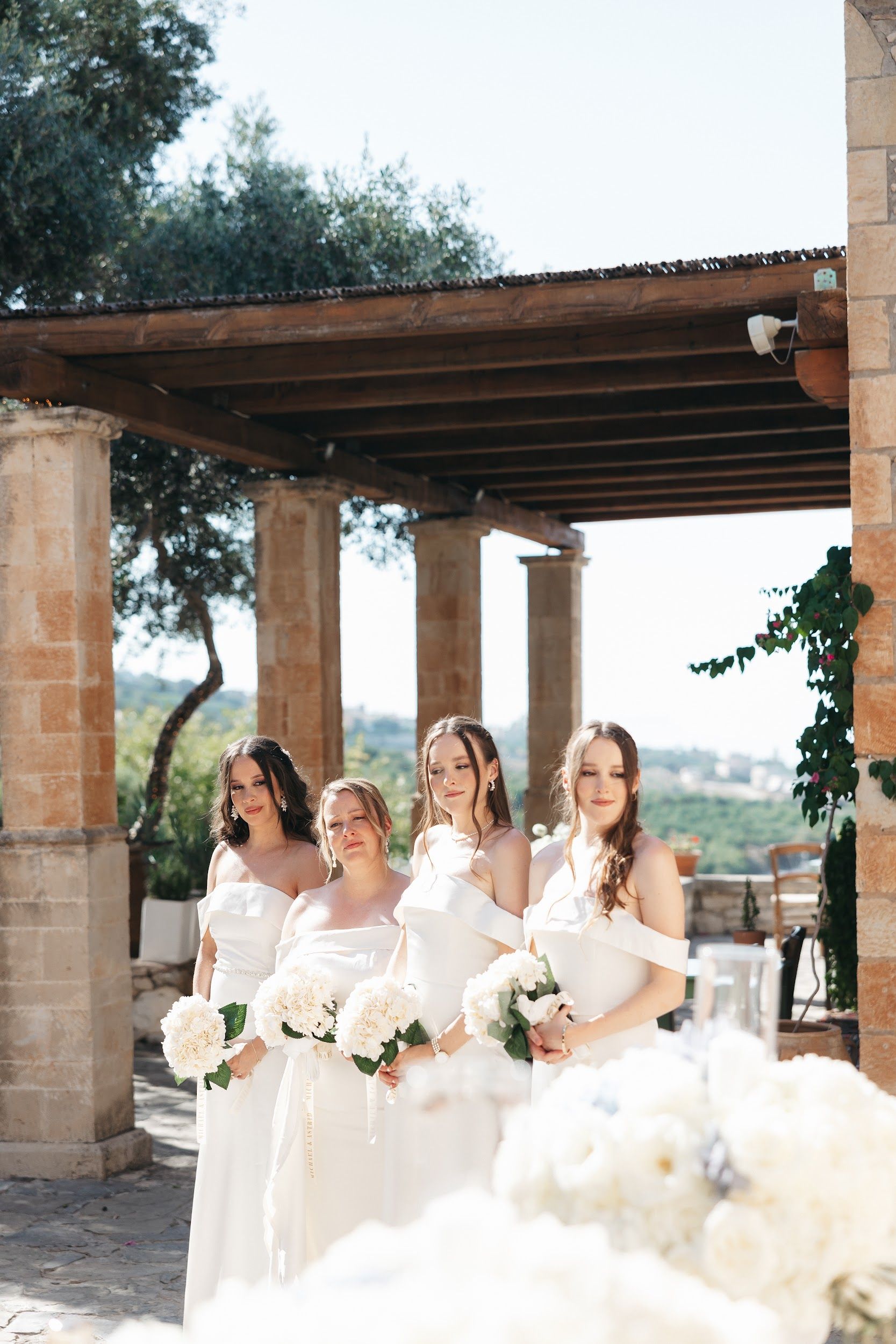 Four bridesmaids in matching white off-the-shoulder gowns stand side by side, each holding a round bouquet of white hydrangea-like blooms with green leaves, under a rustic timber pergola supported by sandstone columns at Agreco Farm, a wedding venue in Greece. Warm sunlight falls across the stone, and an olive tree and a soft view of green hills open up behind them. Their gentle, composed expressions set a calm, elegant tone for this wedding flower display.