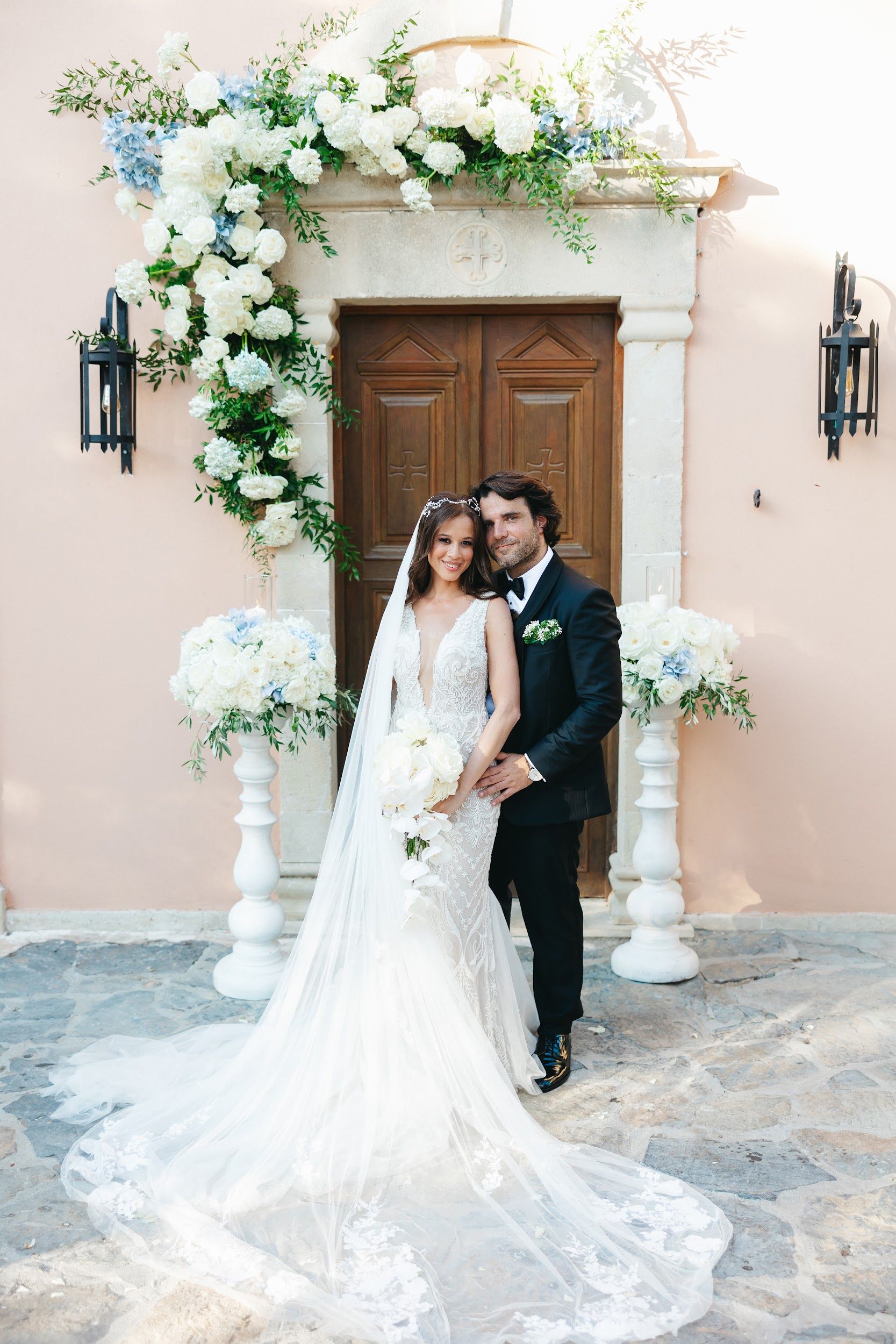 Wedding flowers at Agreco Farm in Greece: a lush arch of white garden roses, white hydrangeas and pale blue hydrangeas with trailing greenery frames a carved wooden door set in a blush pink wall. Two white pedestal urns of roses and hydrangeas flank the couple, who stand close together in the soft daylight: the bride in a lace gown with a long veil holds a white orchid bouquet, and the groom wears a black tuxedo with a small white buttonhole.