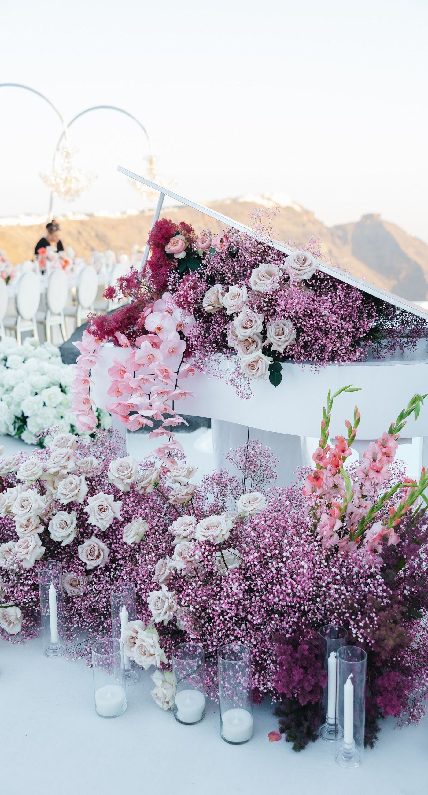 Armita & Azad - A vision of elegance unfolds under a canopy of ethereal blue foliage, accented with white blooms, creating a stunning backdrop for a couple standing in harmonious contrast. Their attire, a dance of monochrome elegance, complements the serene ambiance, while the azure sky and distant cliffs whisper of a hidden paradise. The floor is adorned with cascading white roses, framing the scene with pure grace. The location, with its tranquil vibes and breathtaking views, enhances the overall aesthetic, showcasing a perfect blend of nature and sophistication. This breathtaking display captures the essence of a dreamlike celebration, elevating the floral artistry to new heights. - Photo 5