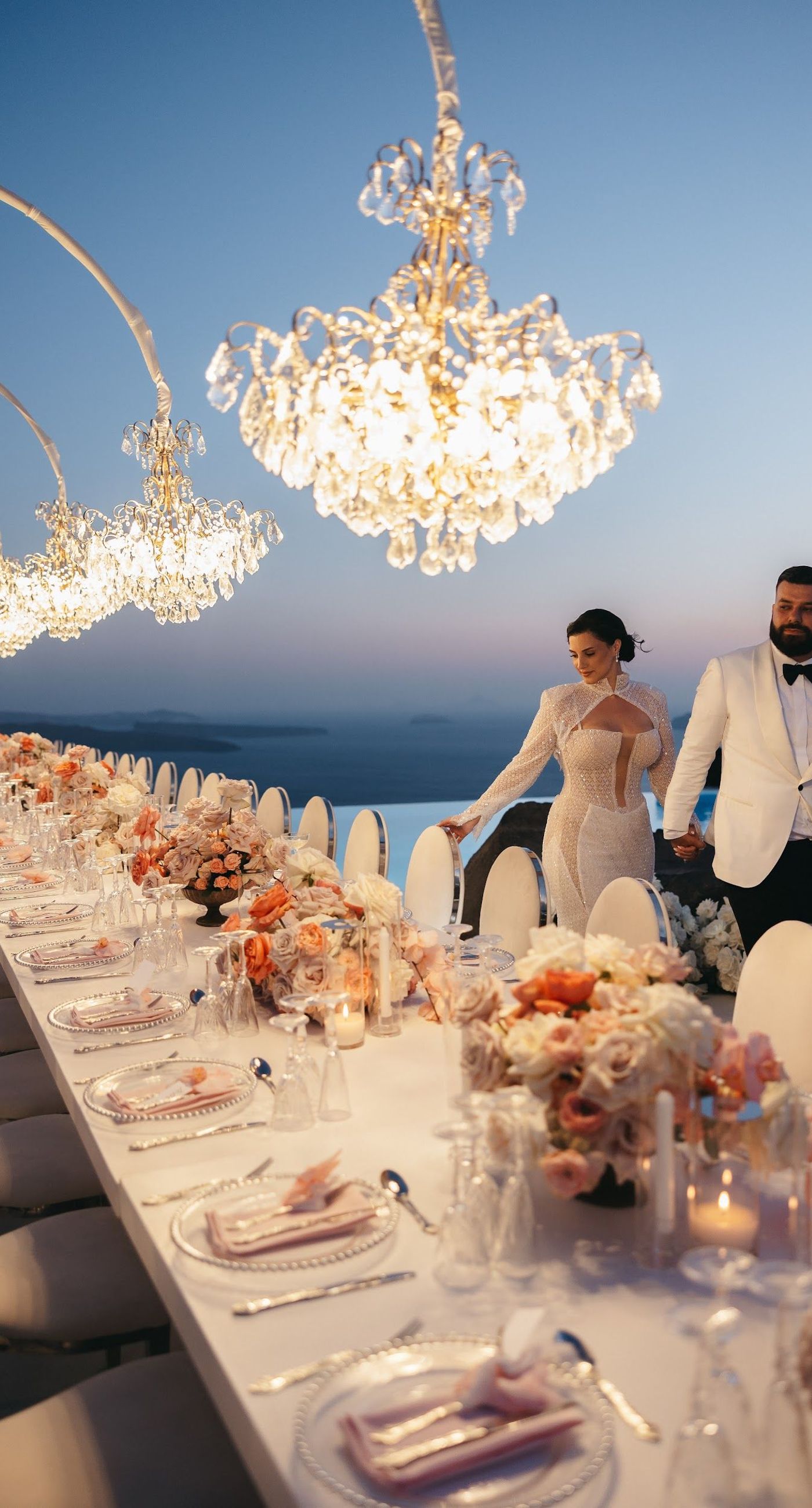 Armita & Azad - A vision of elegance unfolds under a canopy of ethereal blue foliage, accented with white blooms, creating a stunning backdrop for a couple standing in harmonious contrast. Their attire, a dance of monochrome elegance, complements the serene ambiance, while the azure sky and distant cliffs whisper of a hidden paradise. The floor is adorned with cascading white roses, framing the scene with pure grace. The location, with its tranquil vibes and breathtaking views, enhances the overall aesthetic, showcasing a perfect blend of nature and sophistication. This breathtaking display captures the essence of a dreamlike celebration, elevating the floral artistry to new heights. - Photo 9