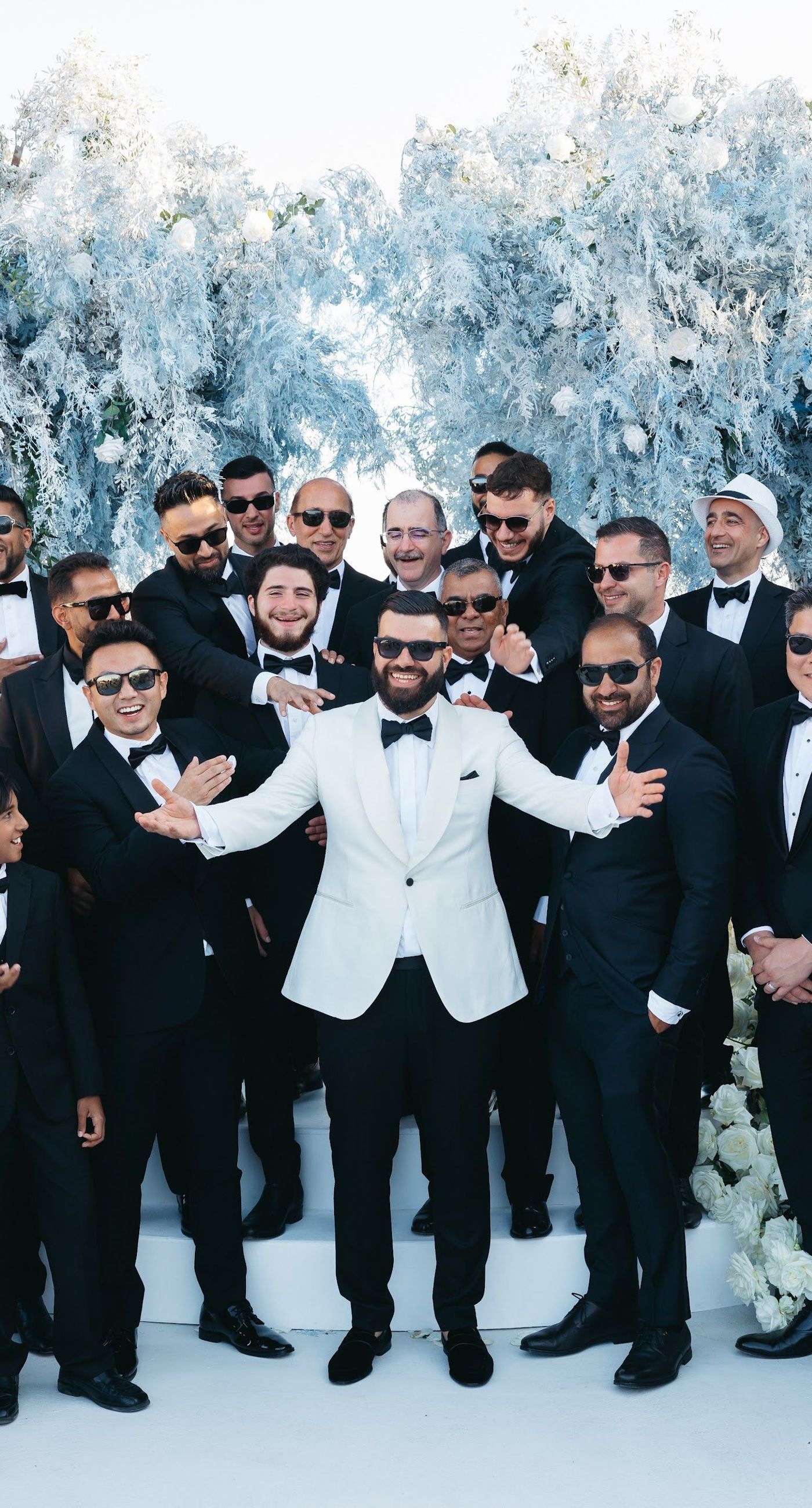 Armita & Azad - A vision of elegance unfolds under a canopy of ethereal blue foliage, accented with white blooms, creating a stunning backdrop for a couple standing in harmonious contrast. Their attire, a dance of monochrome elegance, complements the serene ambiance, while the azure sky and distant cliffs whisper of a hidden paradise. The floor is adorned with cascading white roses, framing the scene with pure grace. The location, with its tranquil vibes and breathtaking views, enhances the overall aesthetic, showcasing a perfect blend of nature and sophistication. This breathtaking display captures the essence of a dreamlike celebration, elevating the floral artistry to new heights. - Photo 4