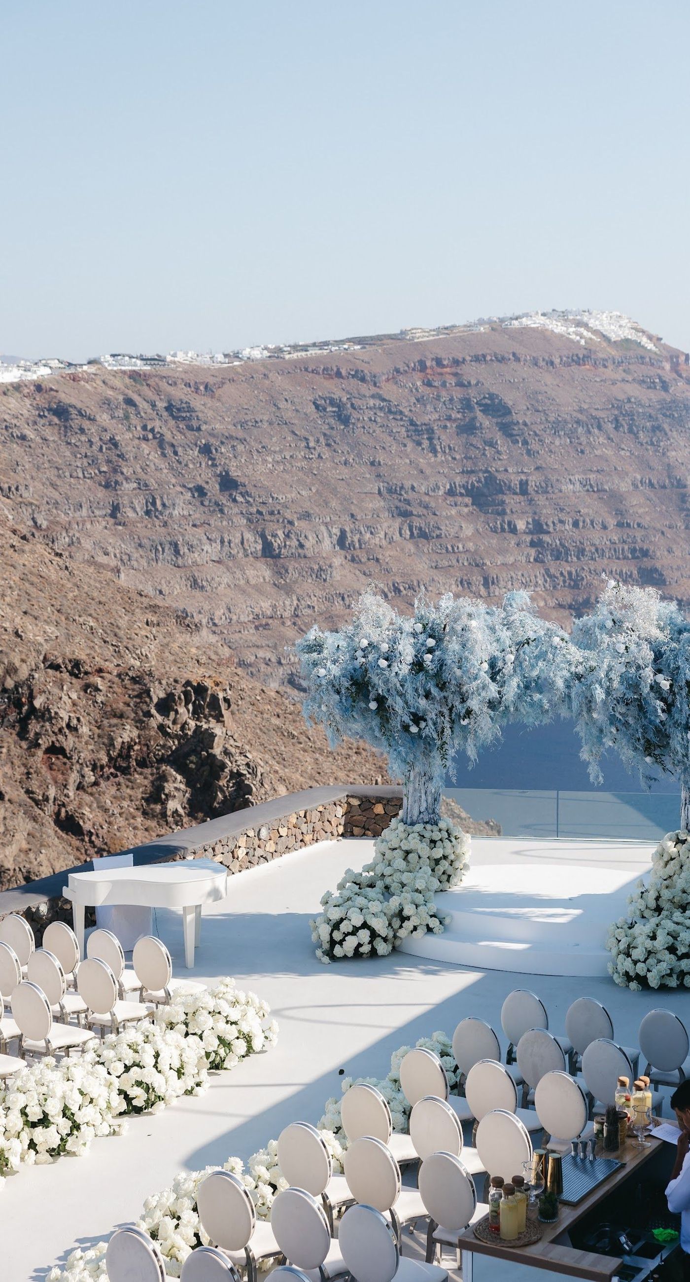 Armita & Azad - A vision of elegance unfolds under a canopy of ethereal blue foliage, accented with white blooms, creating a stunning backdrop for a couple standing in harmonious contrast. Their attire, a dance of monochrome elegance, complements the serene ambiance, while the azure sky and distant cliffs whisper of a hidden paradise. The floor is adorned with cascading white roses, framing the scene with pure grace. The location, with its tranquil vibes and breathtaking views, enhances the overall aesthetic, showcasing a perfect blend of nature and sophistication. This breathtaking display captures the essence of a dreamlike celebration, elevating the floral artistry to new heights. - Photo 1