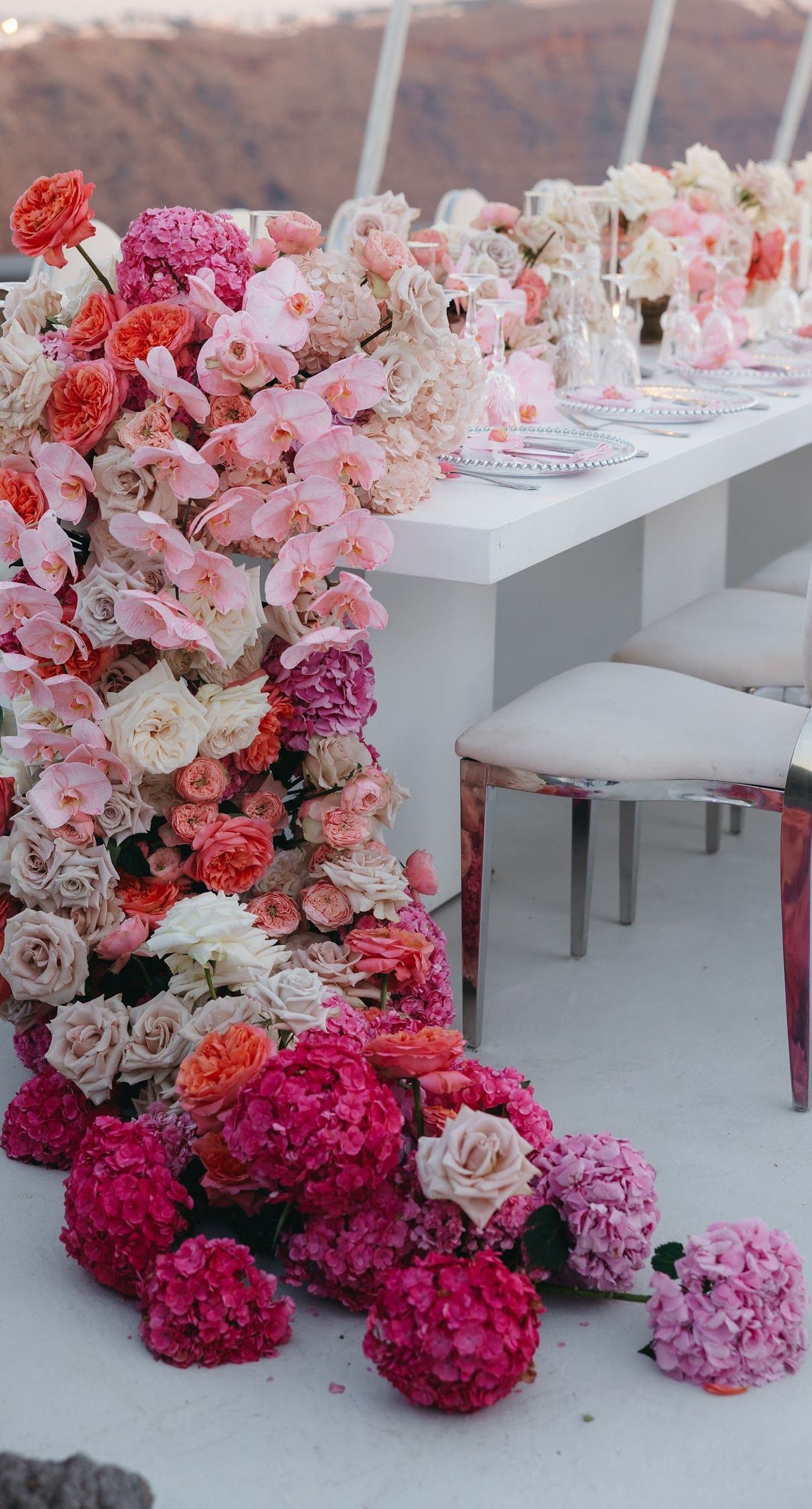 Armita & Azad - A vision of elegance unfolds under a canopy of ethereal blue foliage, accented with white blooms, creating a stunning backdrop for a couple standing in harmonious contrast. Their attire, a dance of monochrome elegance, complements the serene ambiance, while the azure sky and distant cliffs whisper of a hidden paradise. The floor is adorned with cascading white roses, framing the scene with pure grace. The location, with its tranquil vibes and breathtaking views, enhances the overall aesthetic, showcasing a perfect blend of nature and sophistication. This breathtaking display captures the essence of a dreamlike celebration, elevating the floral artistry to new heights. - Photo 7