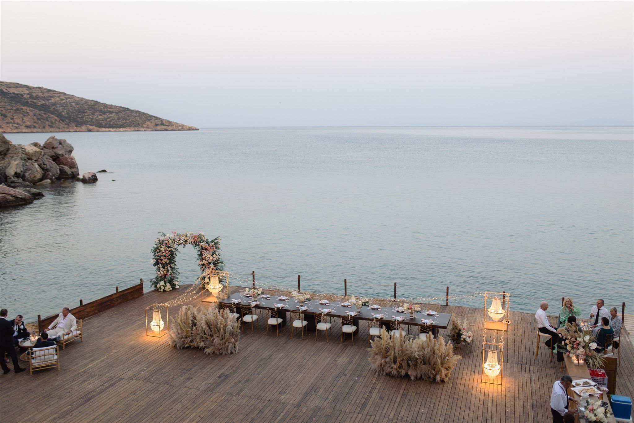 A wooden terrace jutting over calm grey-blue sea at Lazarou Beach in Greece, set for a wedding dinner at dusk. A floral arch of pink and white blooms and greenery stands near the water, with a long dark table below it, laid with white chairs and tall pampas grass clusters at each end. Glowing lanterns and strings of fairy lights line the deck, and a few guests sit relaxed on a lounge to the left beneath a rocky headland.