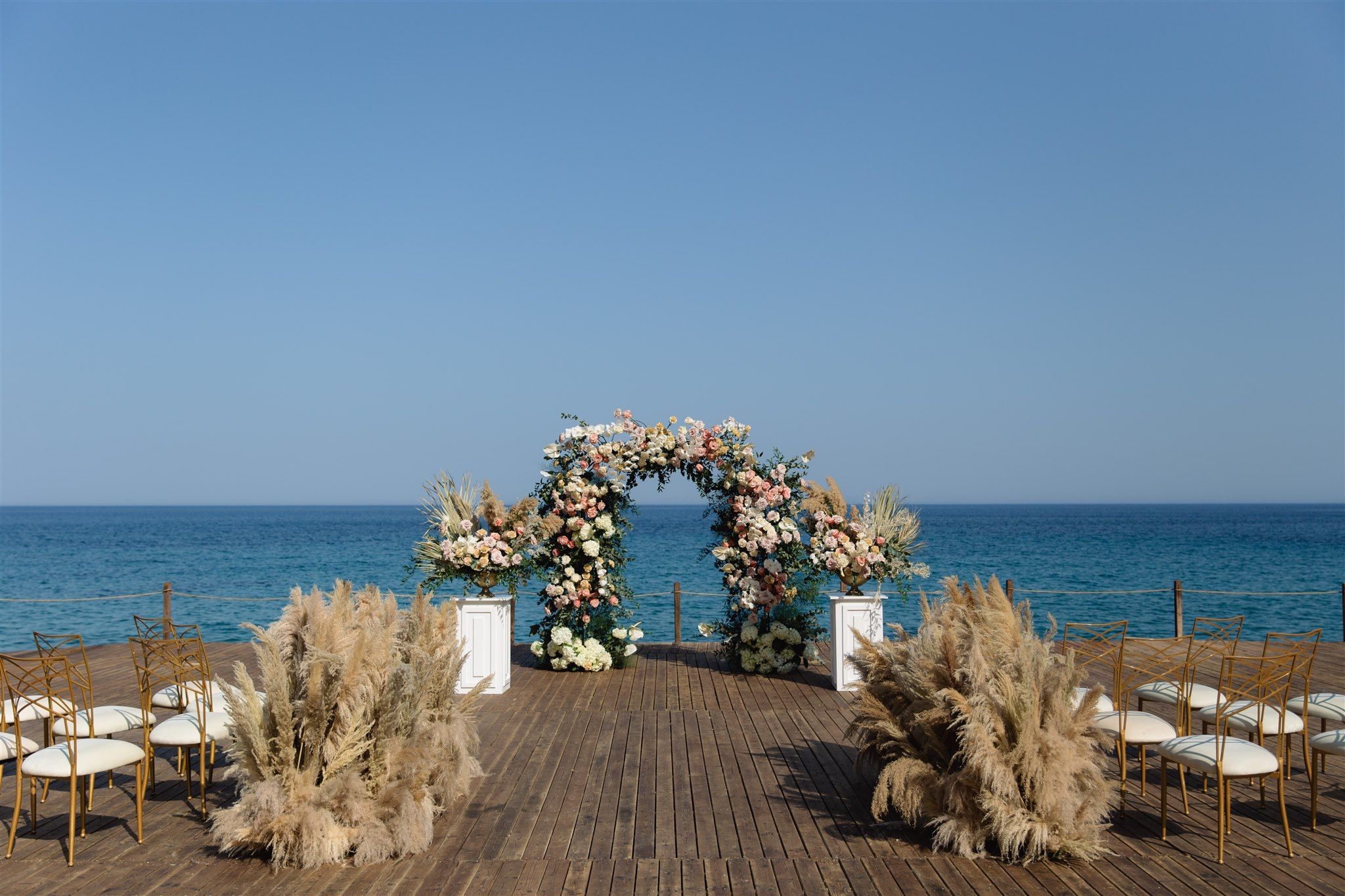 A wedding ceremony arch on a wooden deck overlooking the deep blue Aegean at Lazarou Beach in Greece, woven with blush garden roses, cream hydrangeas, white blooms and trailing green foliage. Two white pedestals hold matching floral urns, and dried pampas grass clusters flank the aisle, which is lined with gold chairs with cream seats. Under a clear, pale sky, the soft pinks and creams stand out against the sea, creating a calm, romantic setting for wedding flowers in Greece.