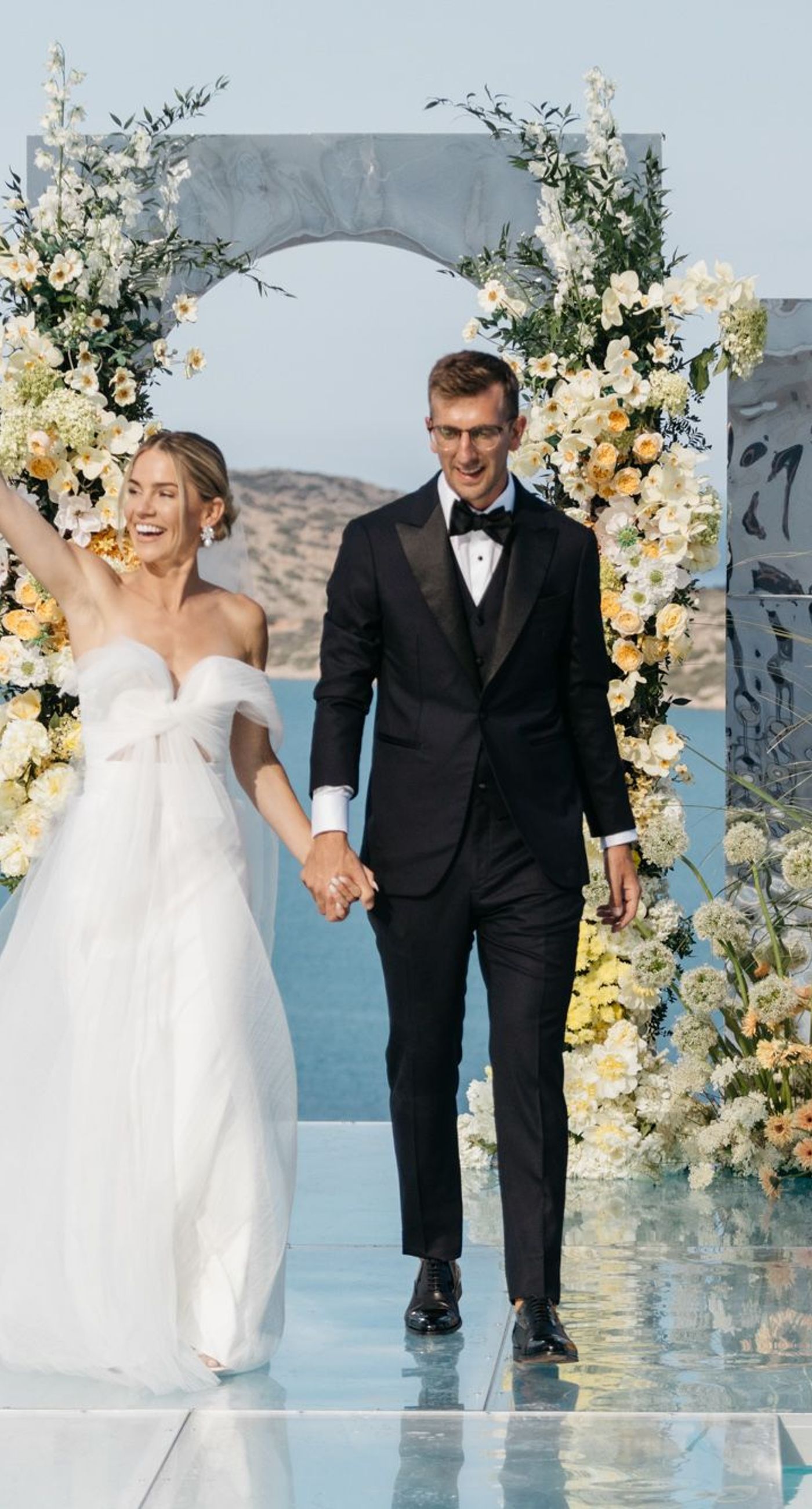 Carla & John - In a sunlit embrace, the bride and groom share a moment of joy amidst a backdrop of elegant floral artistry. The gentle hues of cream, blush, and soft yellow flowers harmonize with lush, verdant greens, creating a serene and timeless palette. The azure sky and distant sea enhance this romantic tableau, adding a sense of boundless possibilities. This composition is a testament to the delicate interplay of nature’s beauty and human emotion, where each petal and leaf is placed with thoughtful precision, embodying the essence of love and celebration. - Photo 5