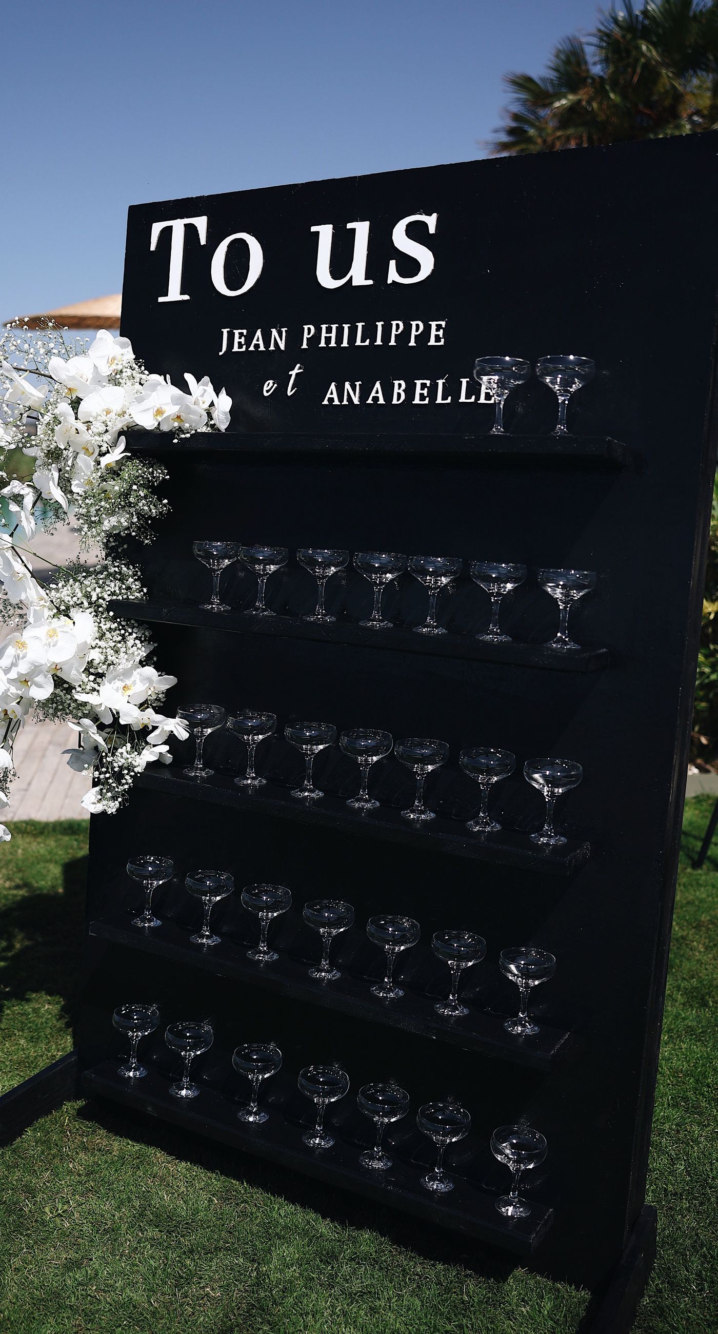 Charlotte & Mateo - In a sunlit scene that marries elegance and joy, a couple strides down an aisle adorned with delicate white petals, while attendees cheerfully shower them with admiration and celebratory petals, embodying a spirit of love and unity. The bride's radiant white gown contrasts beautifully with the subtle greens of the surroundings and the soft blue sky, as the groom's sleek black and white ensemble complements the sophisticated atmosphere. Lush greenery and a graceful arch laced with florals set an enchanting stage, making this moment an exquisite testimony to a florist's craft, capturing both the grandeur and warmth of the occasion. - Photo 8