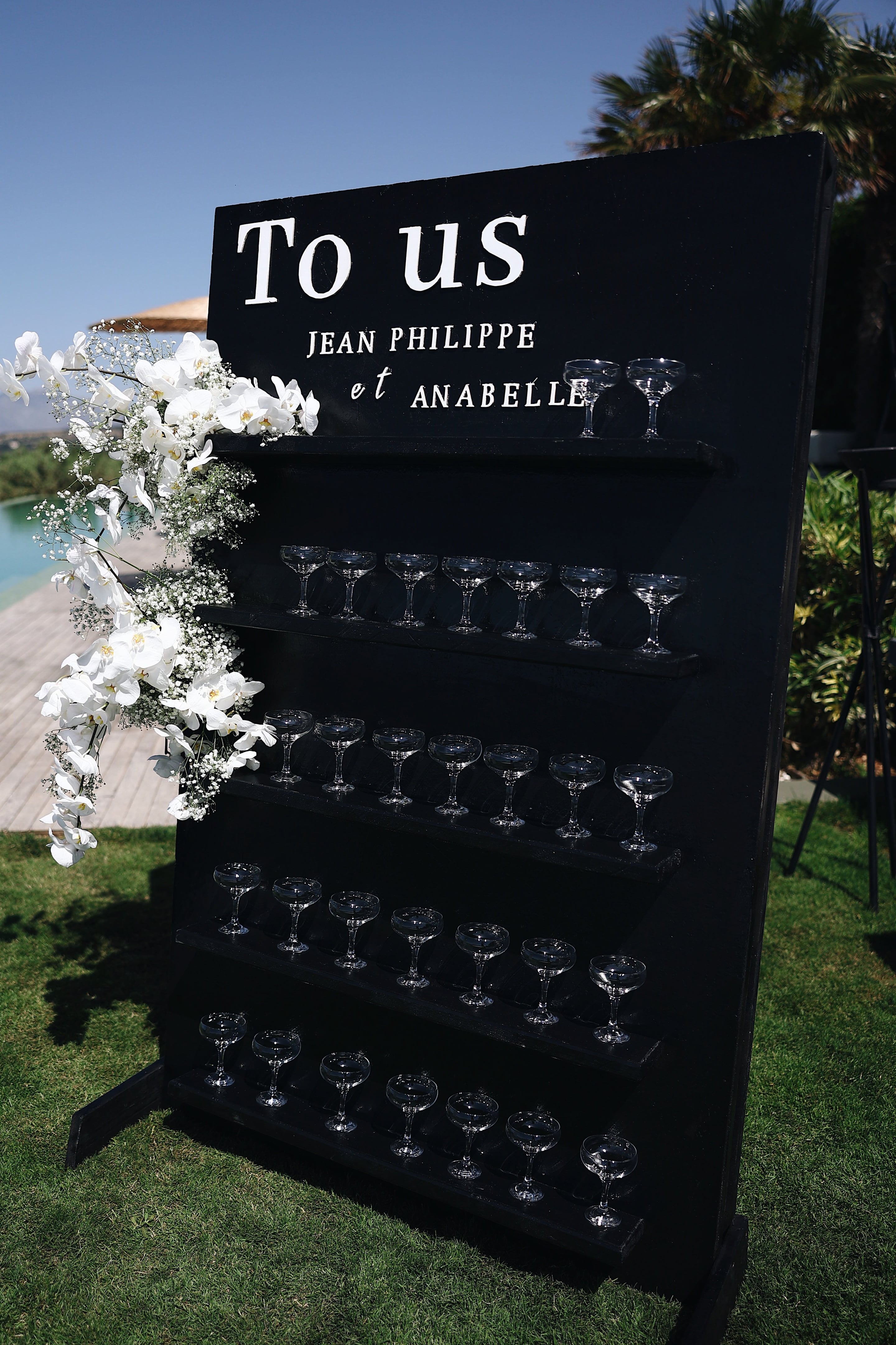 A tall black wooden champagne tower at a wedding reception at Villa Amnalys in Greece, its white lettering reading "To us, Jean Philippe et Anabelle." Rows of empty crystal coupe glasses sit on three tiers beneath the lettering, and a cascade of white phalaenopsis orchids and baby's breath trails down its left edge. Bright sunlight and a clear blue sky frame the scene, with a glimpse of a pool and green foliage behind.