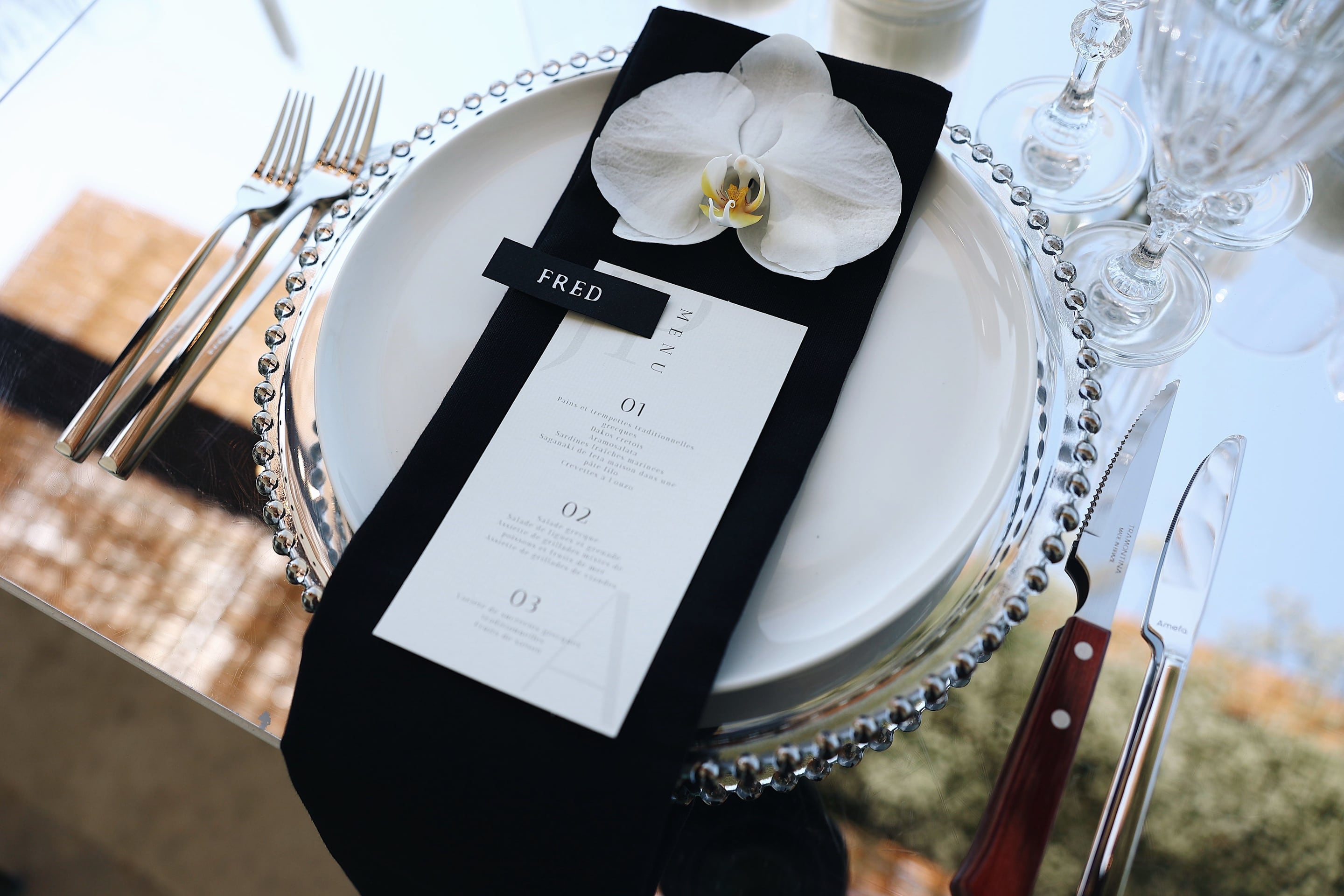 A place setting for a wedding table at Villa Amnalys in Greece, with a single white orchid resting on a folded black napkin. The white charger is edged with a beaded silver rim, and a cream menu card with a black "FRED" name tag sits across the napkin, with silver cutlery and a stemmed glass to the side. The bold black, white and silver palette and the crisp, glossy light give the table a sleek, elegant feel.