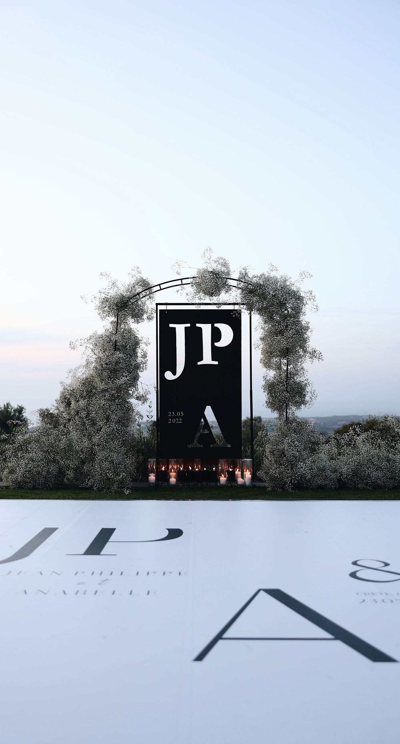 Charlotte & Mateo - In a sunlit scene that marries elegance and joy, a couple strides down an aisle adorned with delicate white petals, while attendees cheerfully shower them with admiration and celebratory petals, embodying a spirit of love and unity. The bride's radiant white gown contrasts beautifully with the subtle greens of the surroundings and the soft blue sky, as the groom's sleek black and white ensemble complements the sophisticated atmosphere. Lush greenery and a graceful arch laced with florals set an enchanting stage, making this moment an exquisite testimony to a florist's craft, capturing both the grandeur and warmth of the occasion. - Photo 6