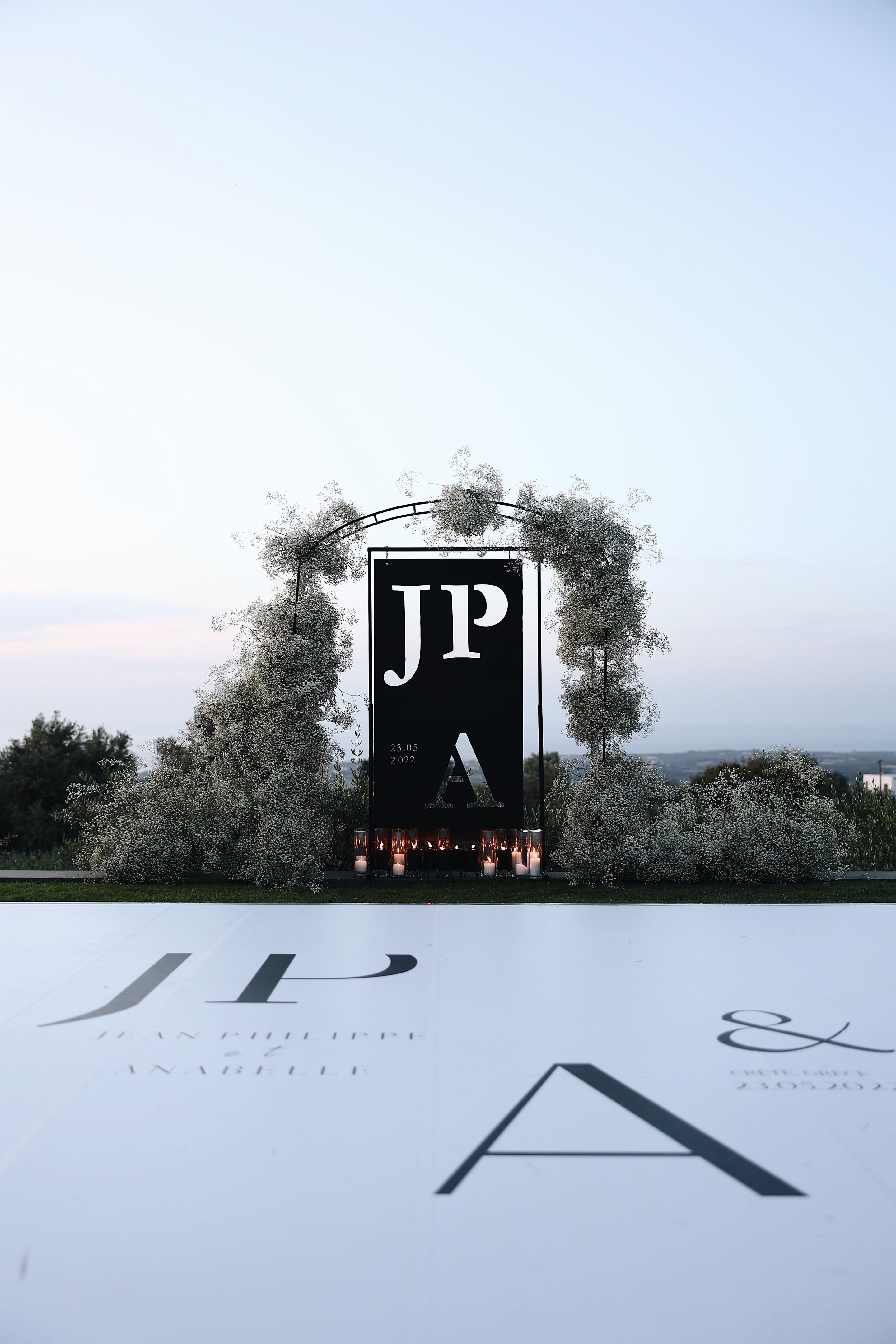 A black monogram panel reading JP, with a large A beneath and the date 23.05 2022 in small white type, stands at the centre of a wedding ceremony at Villa Amnalys in Greece. An arch of dense, cloud-like baby's breath (gypsophila) wraps around the panel, with clusters of candles in glass hurricanes along its base. A white floor in front carries the same initials and an ampersand, and the dusky pale sky and distant hills give the scene a calm, evening feel.