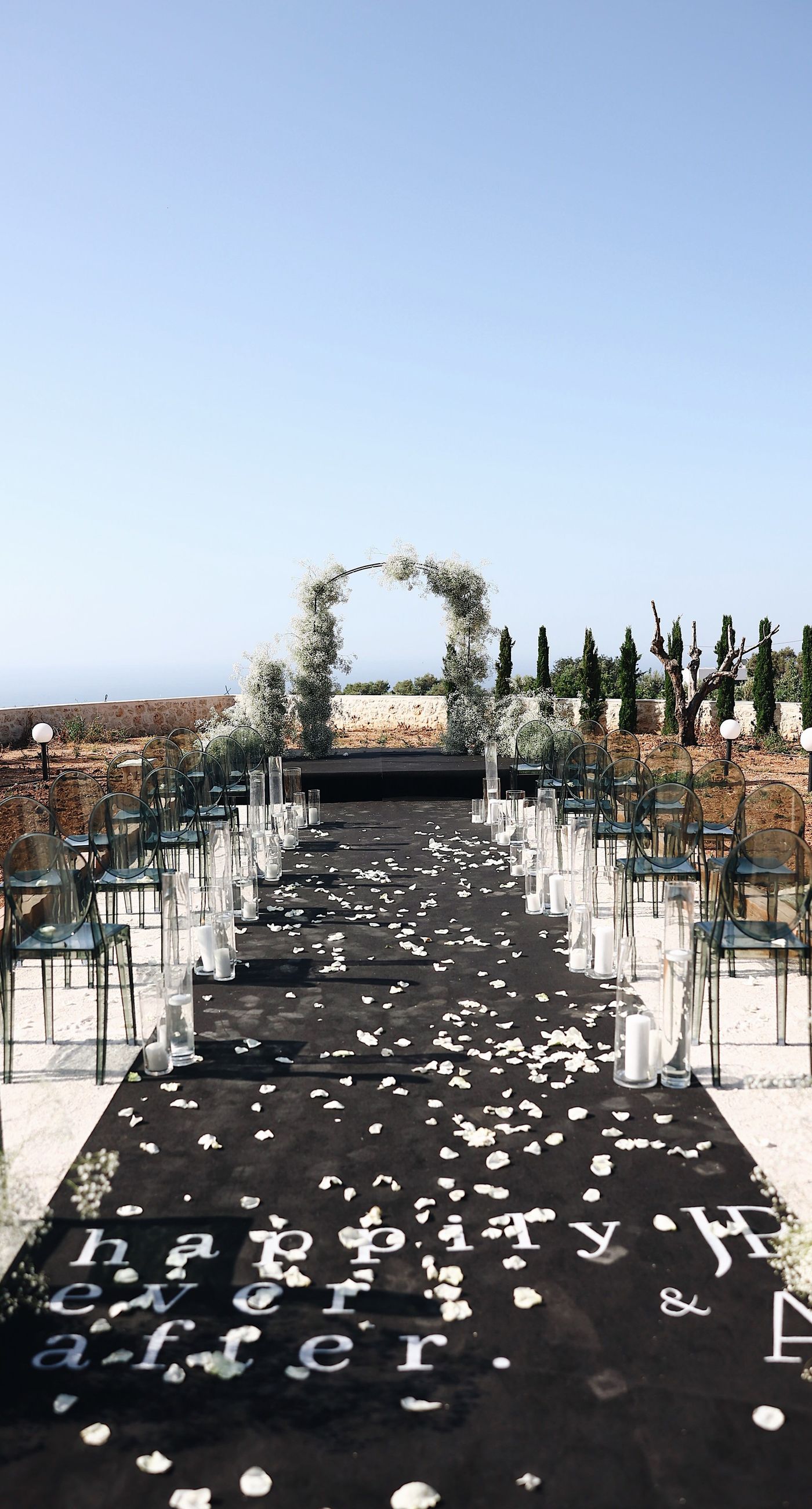 Charlotte & Mateo - In a sunlit scene that marries elegance and joy, a couple strides down an aisle adorned with delicate white petals, while attendees cheerfully shower them with admiration and celebratory petals, embodying a spirit of love and unity. The bride's radiant white gown contrasts beautifully with the subtle greens of the surroundings and the soft blue sky, as the groom's sleek black and white ensemble complements the sophisticated atmosphere. Lush greenery and a graceful arch laced with florals set an enchanting stage, making this moment an exquisite testimony to a florist's craft, capturing both the grandeur and warmth of the occasion. - Photo 1