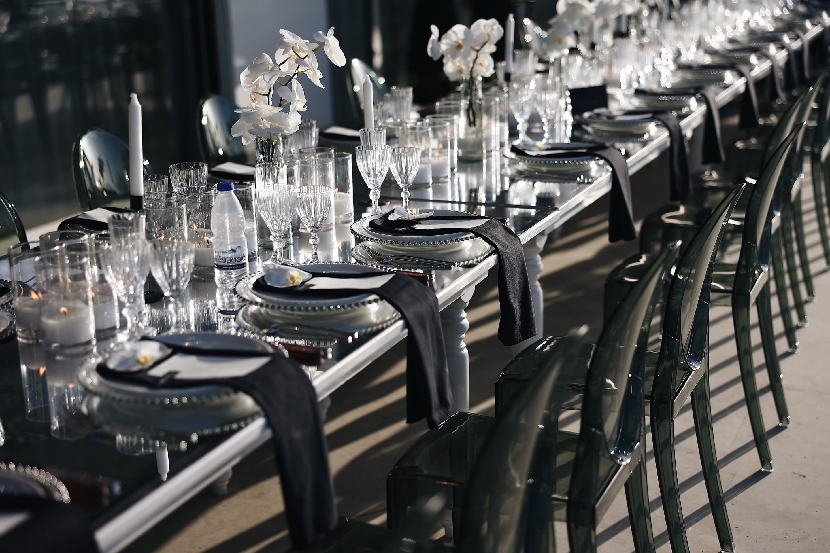 A long banquet table set for a wedding dinner at Villa Amnalys in Greece, running diagonally into the distance on a sunlit terrace. White phalaenopsis orchids rise from slim glass vases between tall white candles, while clear crystal glasses, beaded silver-rimmed plates and black linen napkins line each place setting. Smoky grey acrylic chairs sit along the edge, casting soft striped shadows across the stone floor.