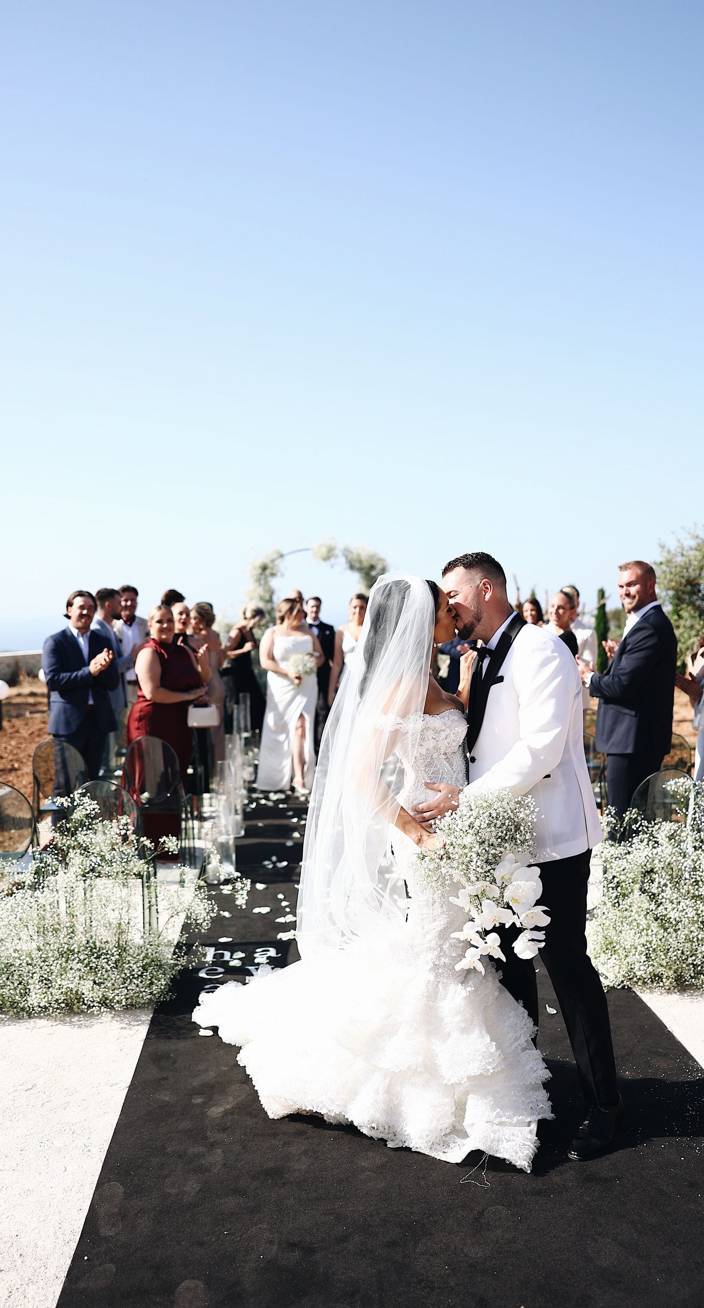 Charlotte & Mateo - In a sunlit scene that marries elegance and joy, a couple strides down an aisle adorned with delicate white petals, while attendees cheerfully shower them with admiration and celebratory petals, embodying a spirit of love and unity. The bride's radiant white gown contrasts beautifully with the subtle greens of the surroundings and the soft blue sky, as the groom's sleek black and white ensemble complements the sophisticated atmosphere. Lush greenery and a graceful arch laced with florals set an enchanting stage, making this moment an exquisite testimony to a florist's craft, capturing both the grandeur and warmth of the occasion. - Photo 2